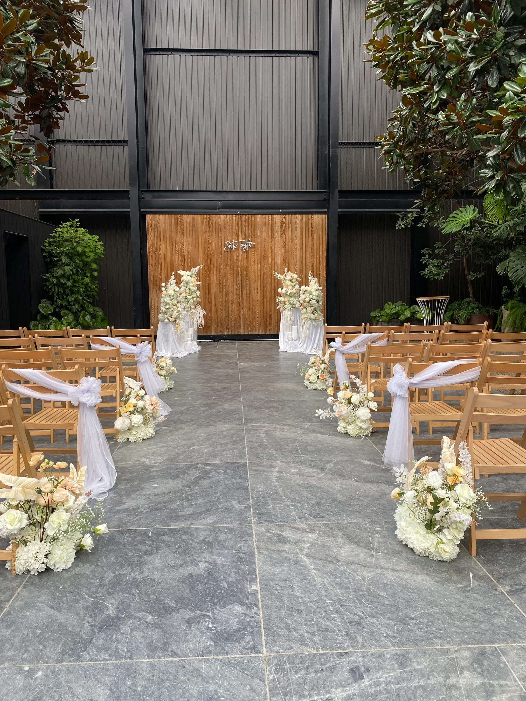 Ceremony styling by Decoration X: Courtyard aisle of timber folding chairs tied with white tulle bows and peach-and-white floral clusters, leading to a timber wall flanked by floral plinths.