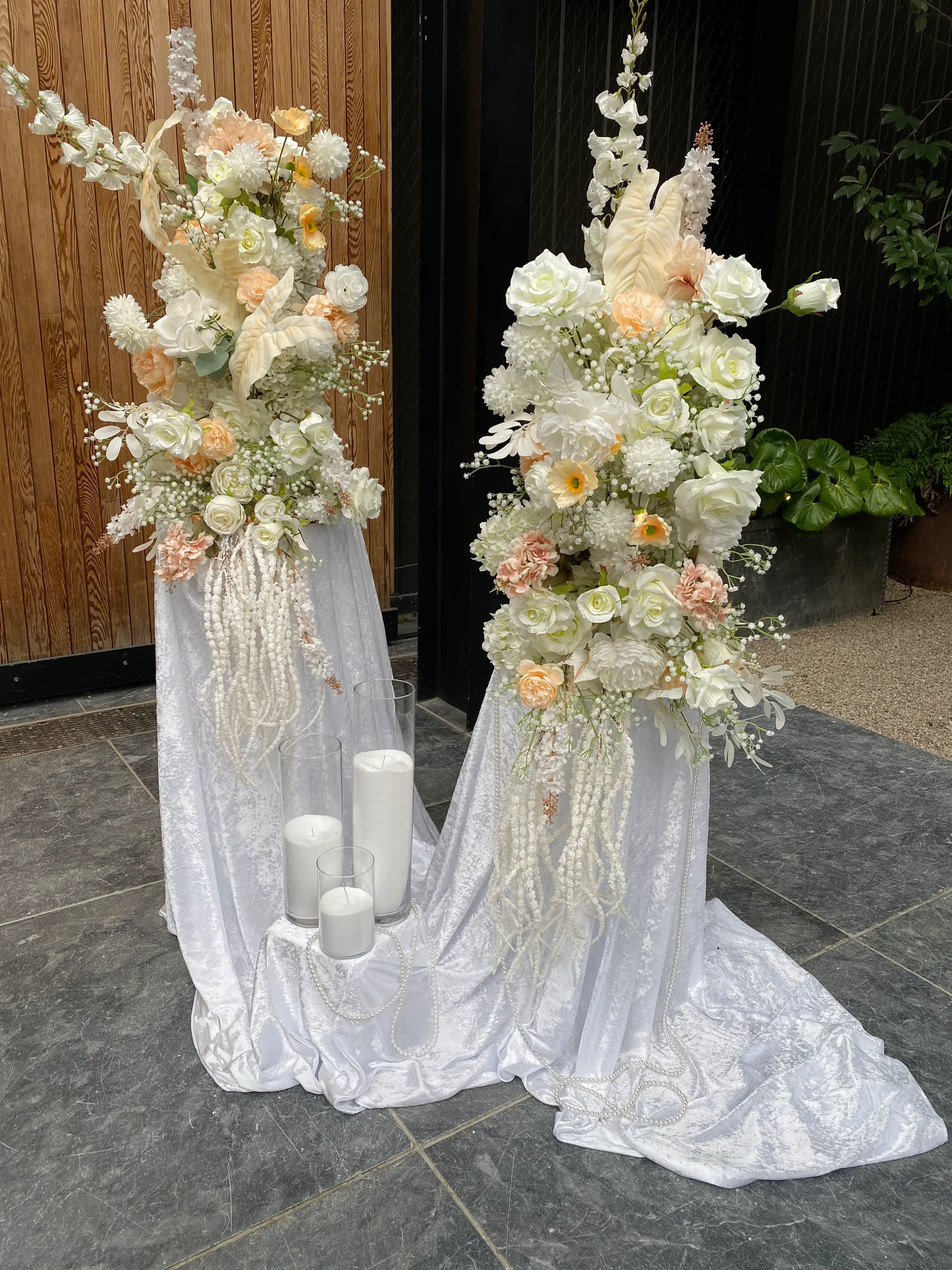 Ceremony styling by Decoration X: Two tall peach, cream and white floral arrangements with anthuriums and hanging amaranthus on white velvet-draped plinths, pillar candles in glass between.