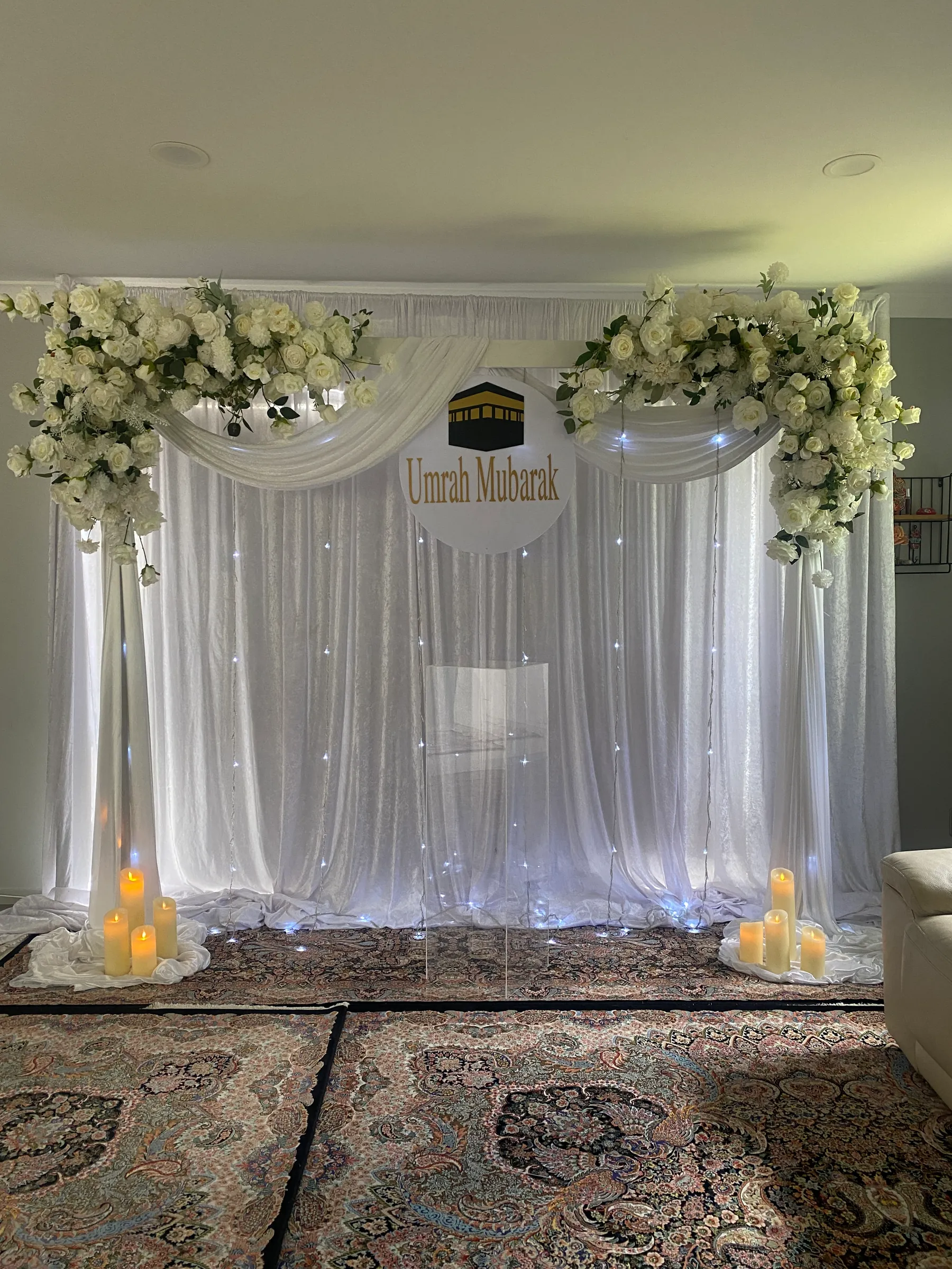 Cultural styling by Decoration X: Fairy-lit white drapes with white rose swags at each corner, a round Umrah Mubarak sign with a Kaaba motif, clear acrylic plinth and candles on Persian rugs.