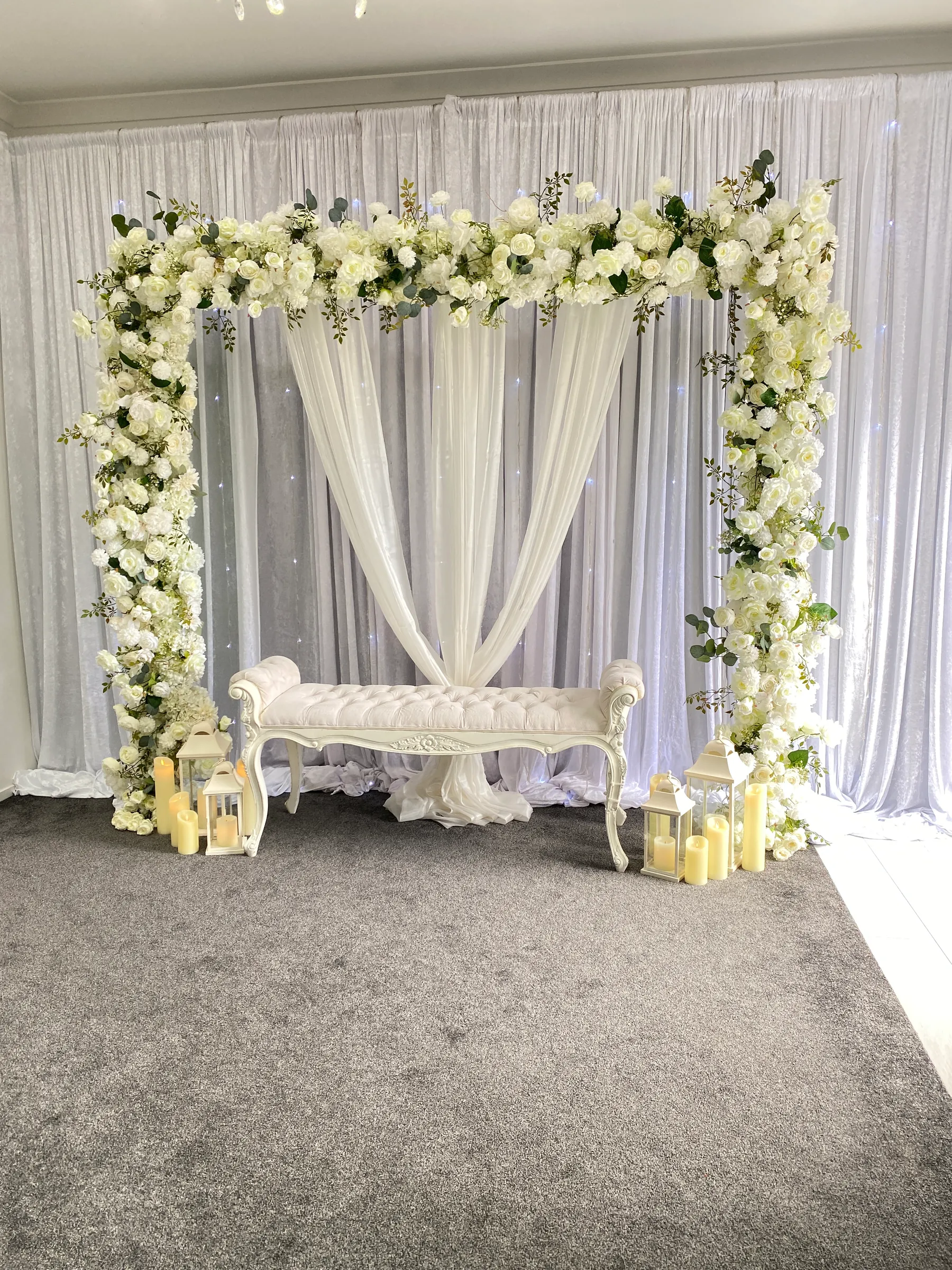 Stage styling by Decoration X: White rose square arch over crossed ivory drapes and a tufted white bench, lanterns and pillar candles at its feet, fairy-lit white drapes behind.