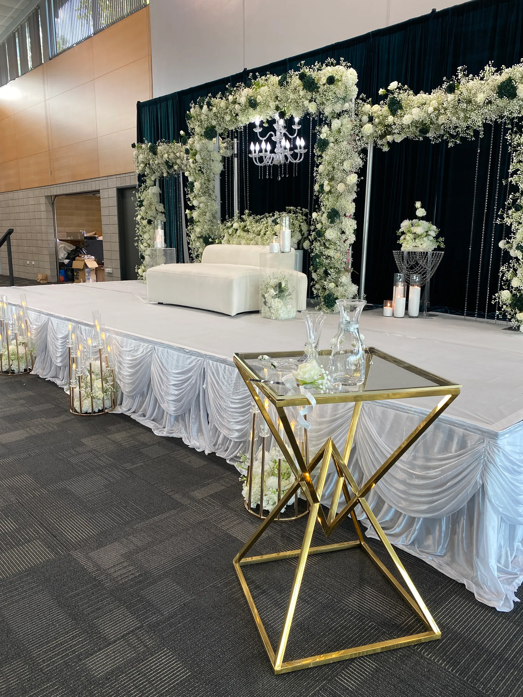 Stage styling by Decoration X: Gold geometric side table with glass carafes before a white-skirted stage, white sofa, gypsophila and rose arches and a chandelier on black drapes.