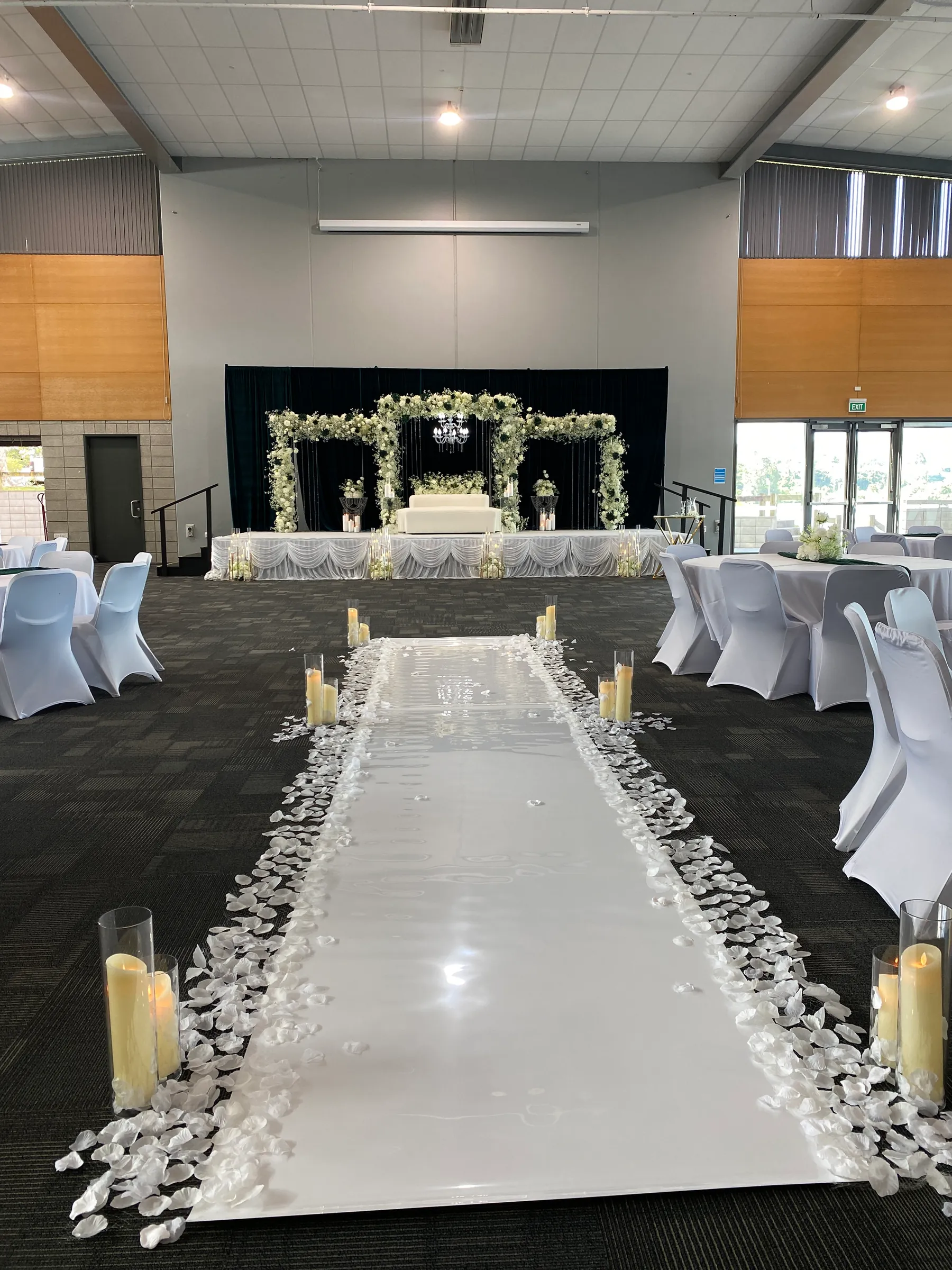 Ceremony styling by Decoration X: Glossy white aisle edged with white petals and pillar candles in glass cylinders, between covered chairs to a floral-arch stage on black drapes.