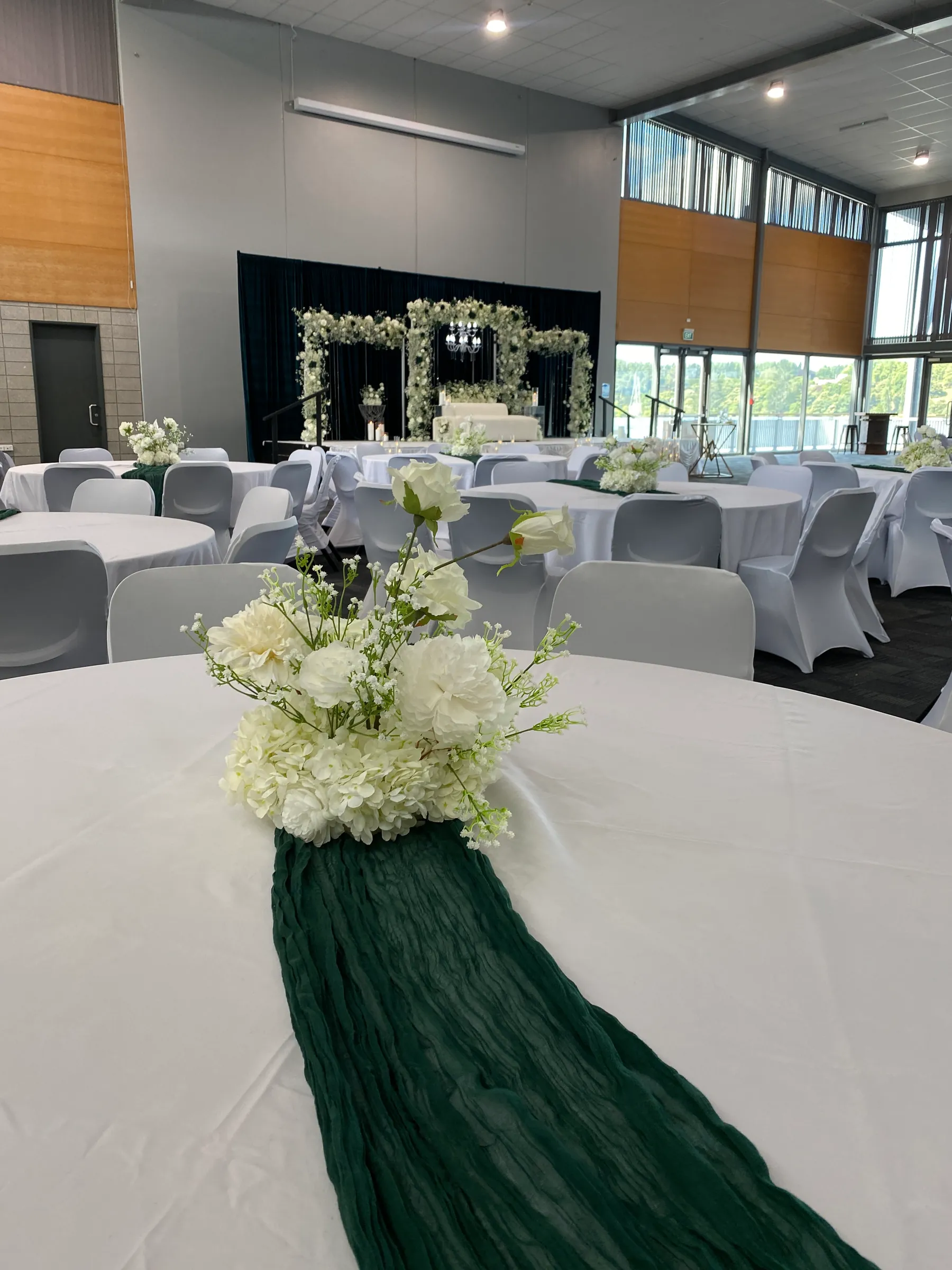 Tables styling by Decoration X: Round table with a white hydrangea and rose centrepiece on a deep-green cheesecloth runner, covered chairs and a floral-arch stage beyond.