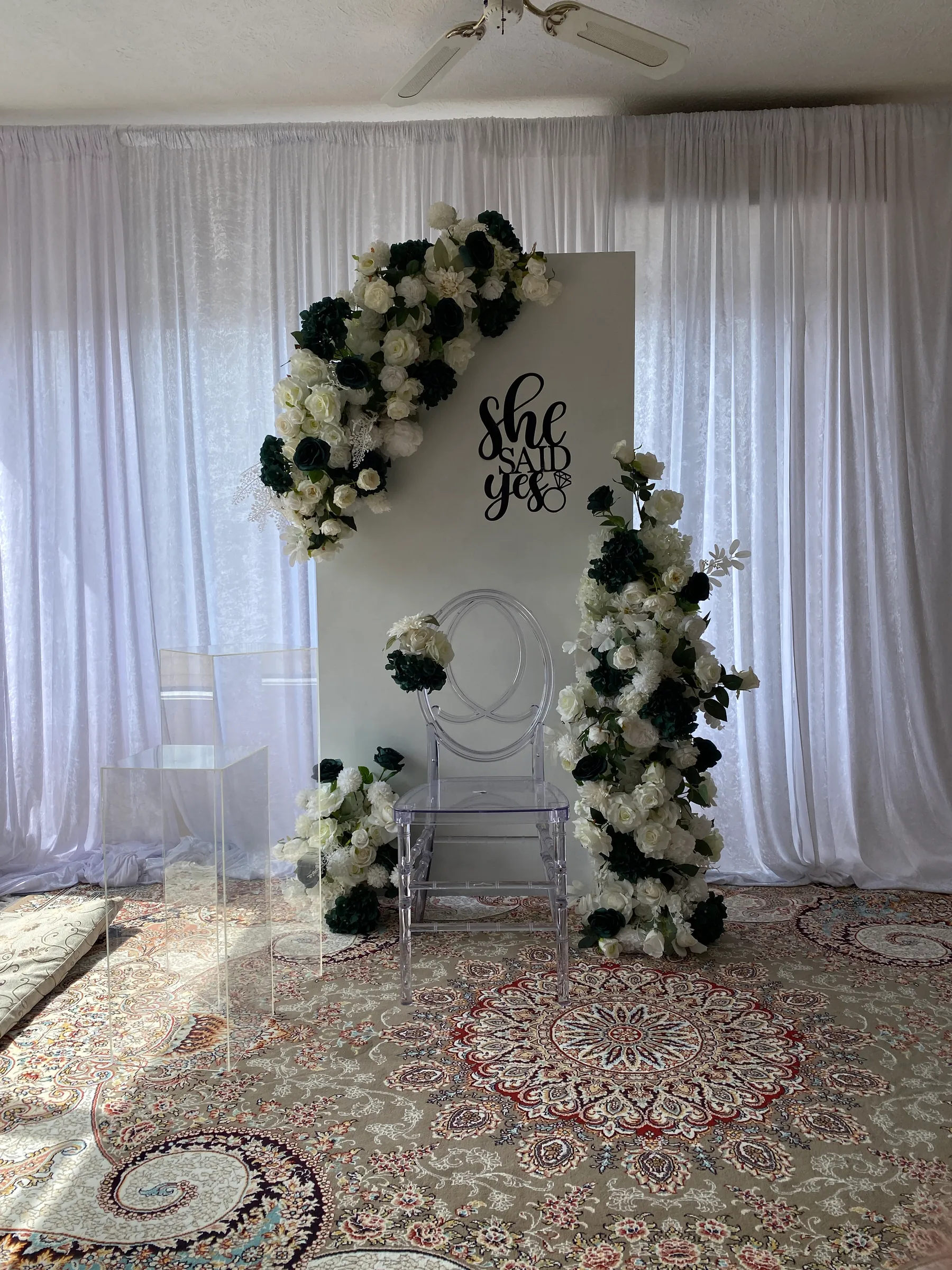 Engagement styling by Decoration X: White panel backdrop with a black script sign and swags of white and deep-green roses, a clear acrylic chair and plinths on a Persian rug.