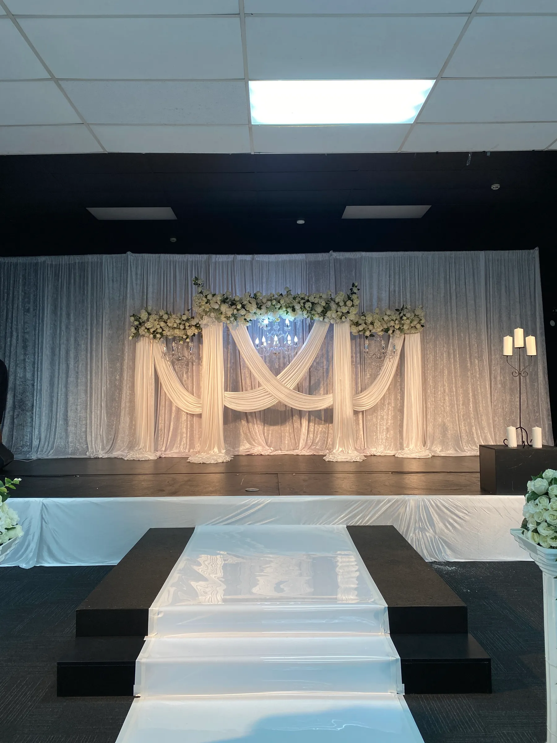 Stage styling by Decoration X: Stage with white crushed-velvet drapes and crossed ivory swags under a white floral garland and chandeliers, a glossy white runner climbing the steps.