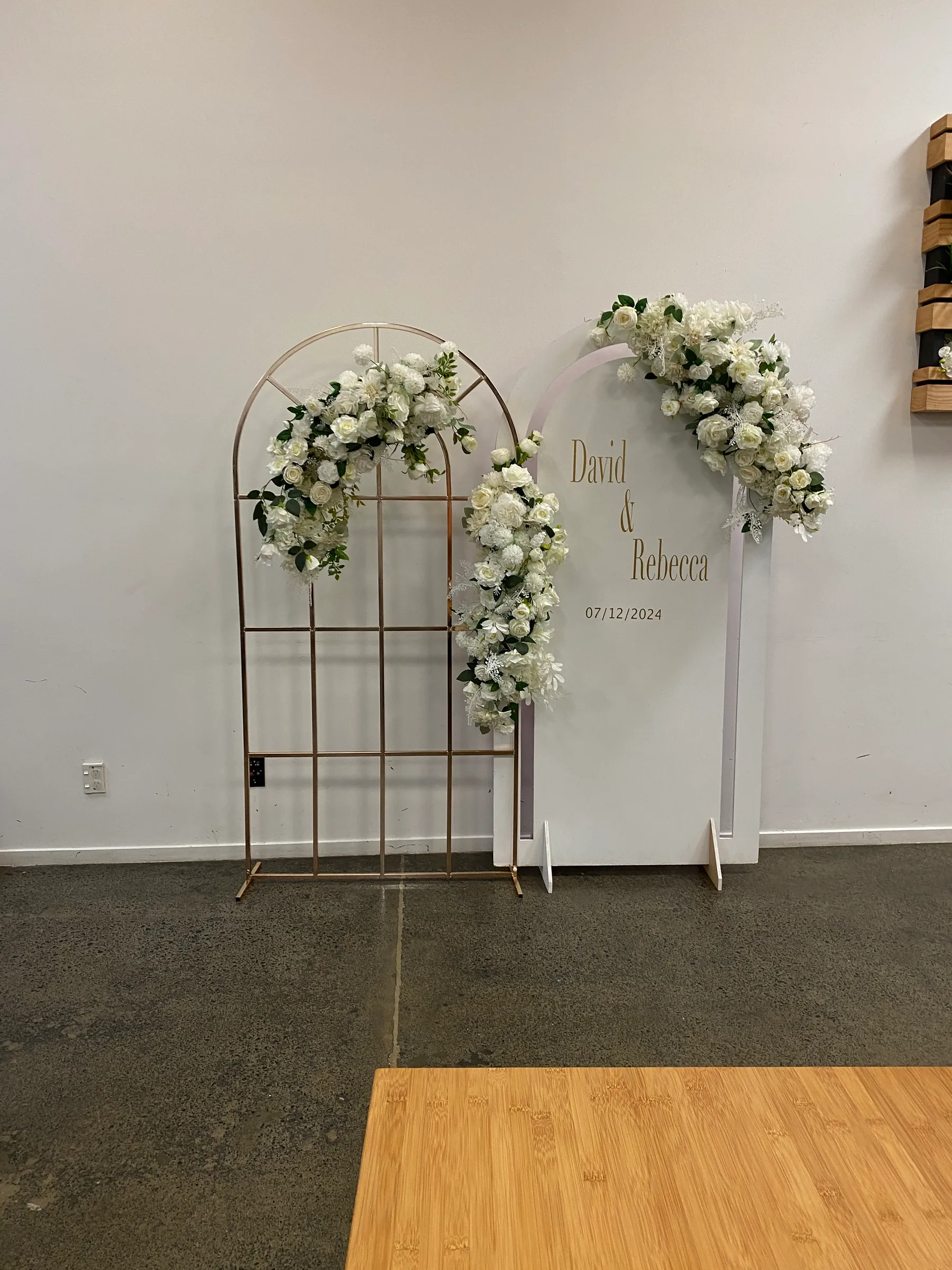 Signage styling by Decoration X: Gold grid arch beside a white acrylic arch name sign, both trailing white roses and greenery, against a plain white wall.