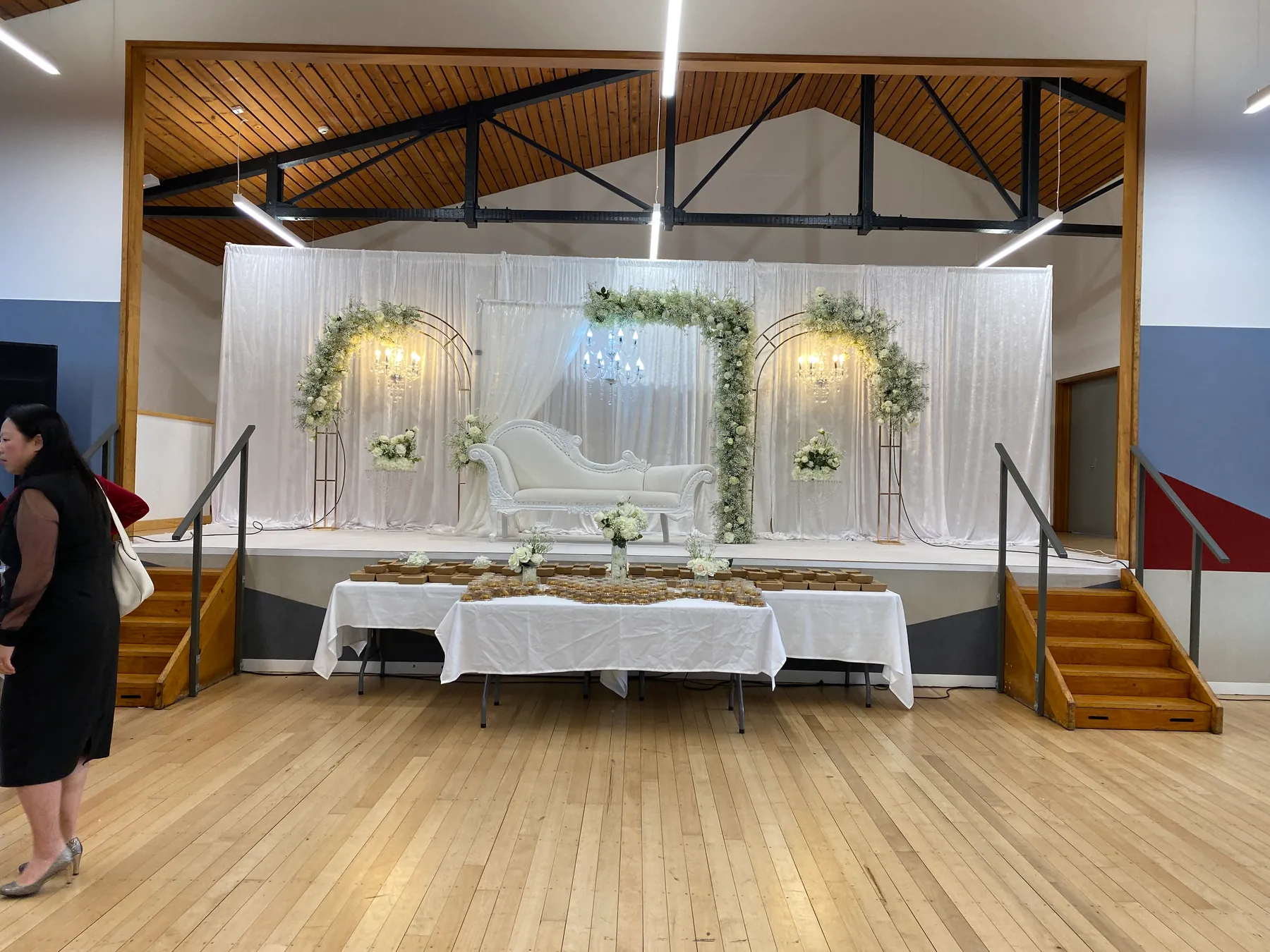 Stage styling by Decoration X: Hall stage in white drapes with three white-and-green floral arches, crystal chandeliers and an ivory carved sofa, a favour table set in front.