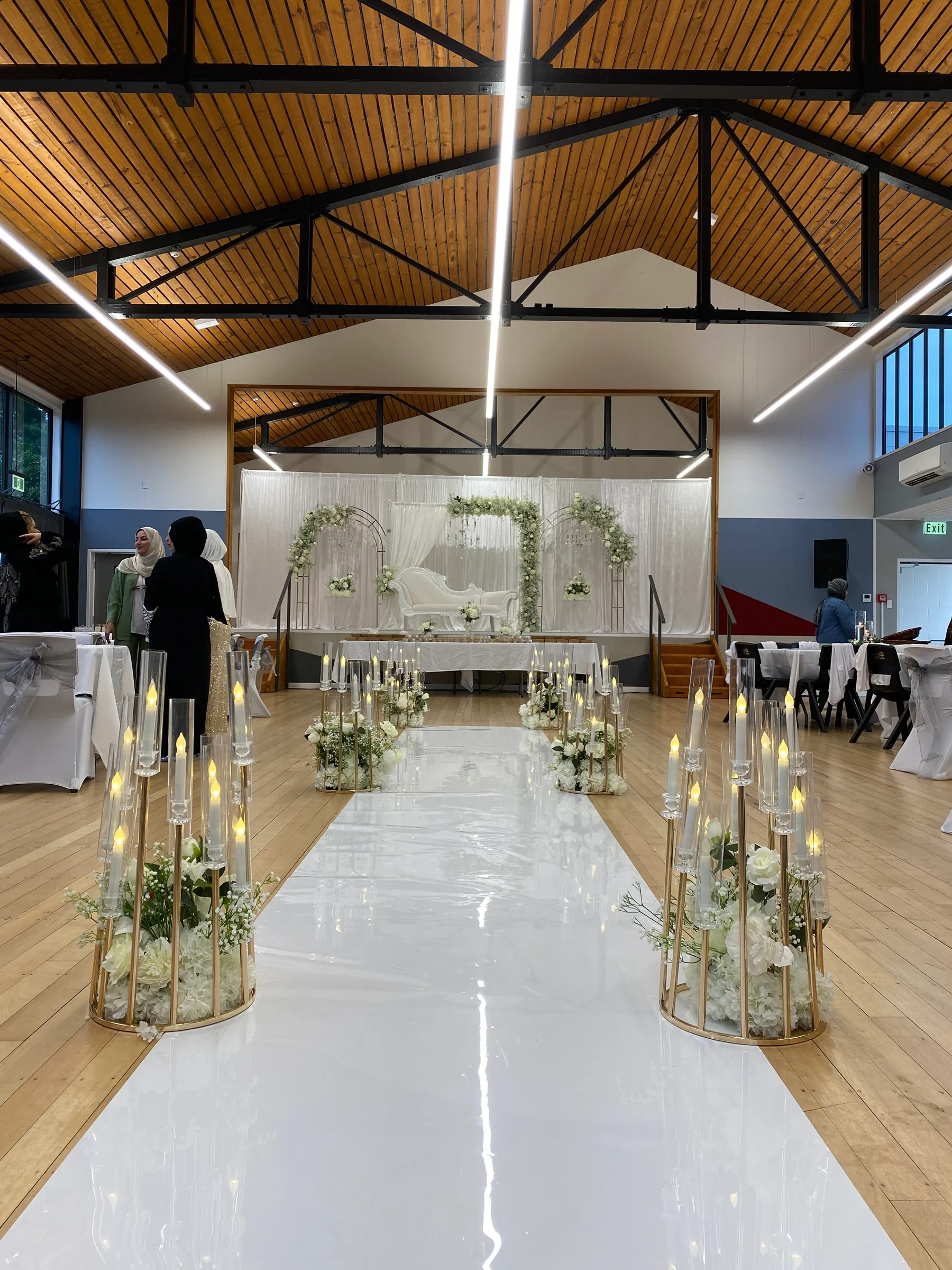 Ceremony styling by Decoration X: Glossy white aisle runner between gold cylinder candelabras with white florals, leading to a draped stage with three greenery arches and an ivory sofa.
