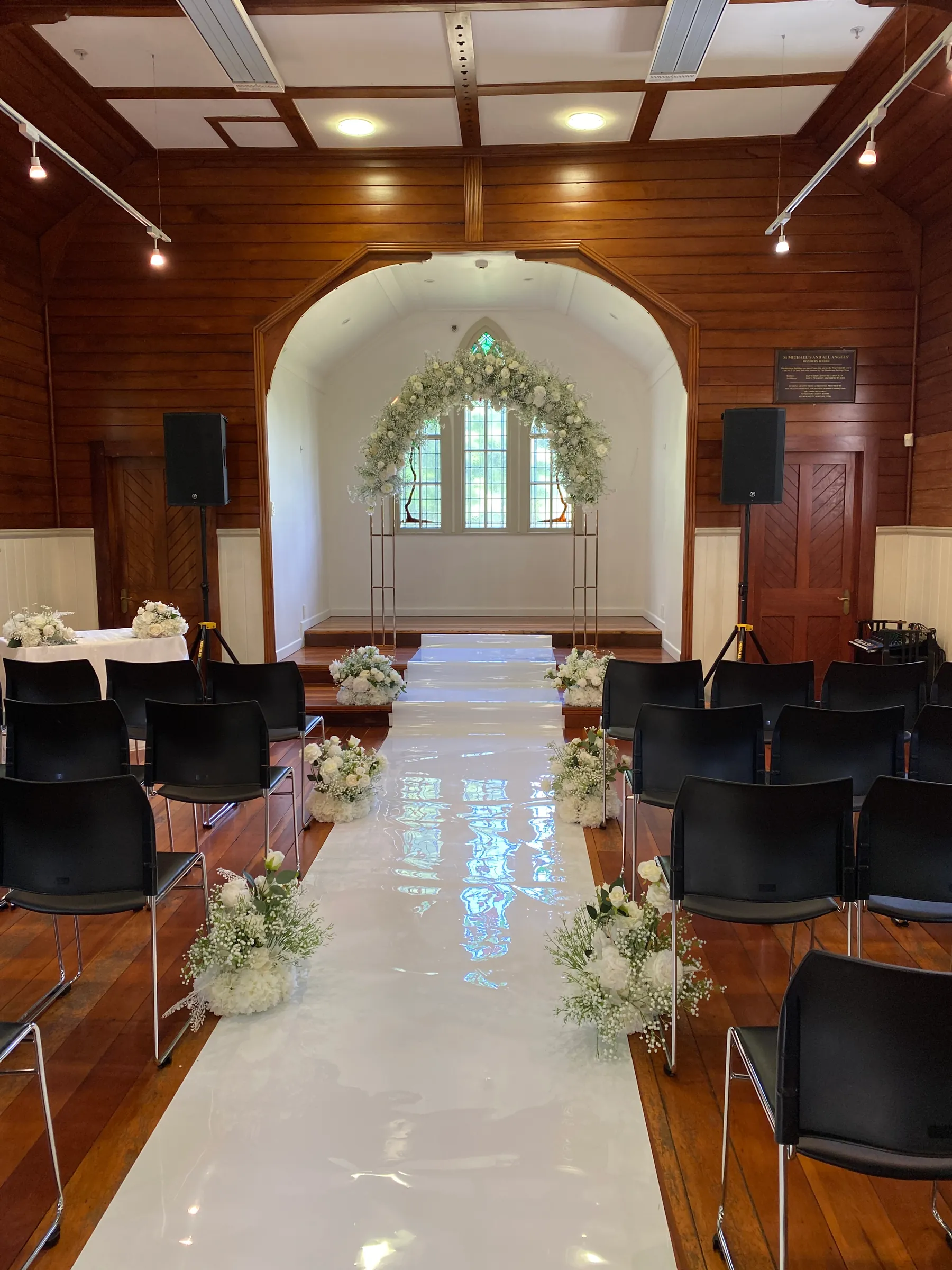 Ceremony styling by Decoration X: Timber-lined chapel with a white gloss aisle runner, gypsophila and hydrangea arrangements along it, a white floral arch before stained-glass windows.