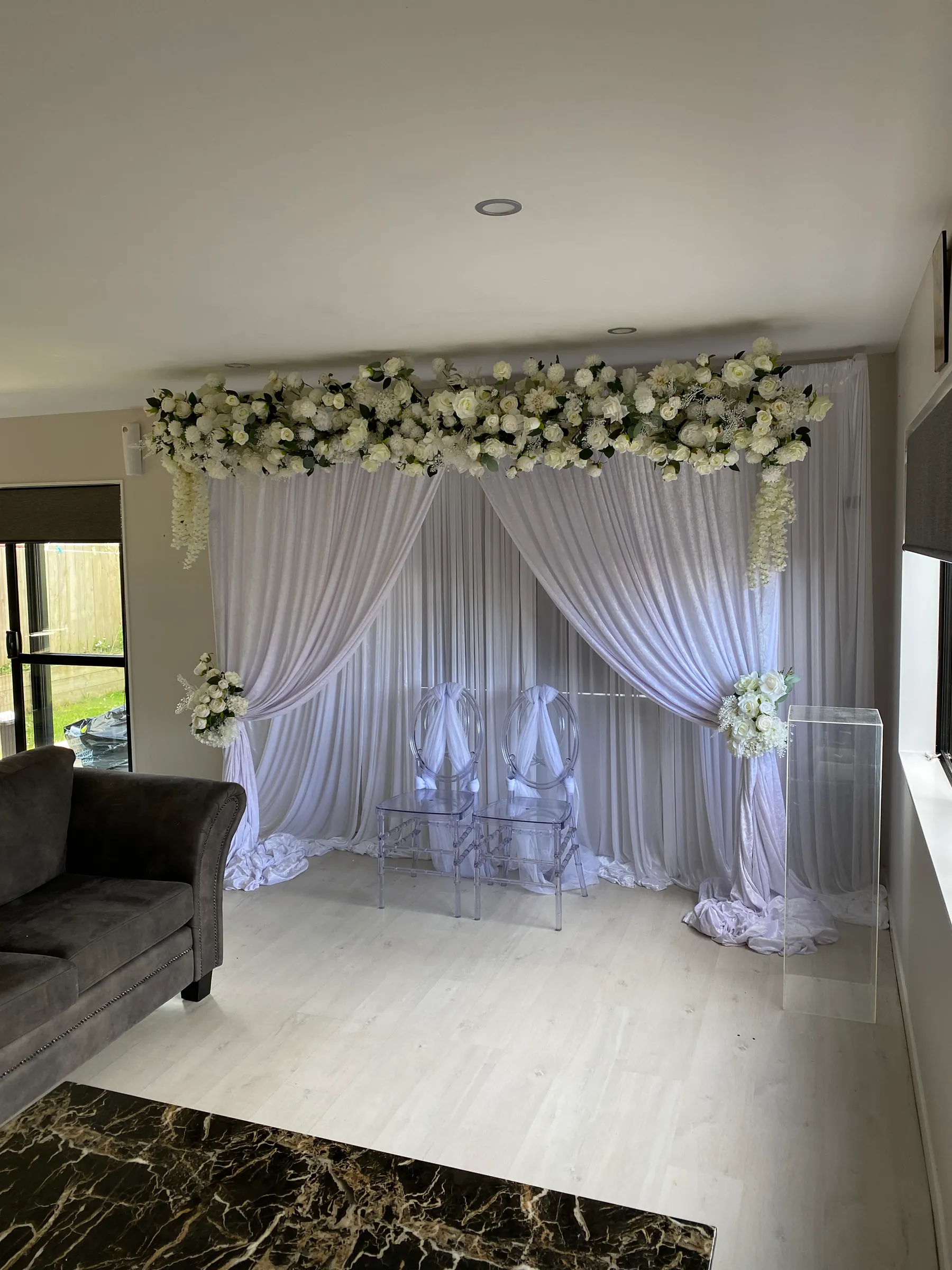 Stage styling by Decoration X: White drape swags under a garland of white roses in a living room, two clear ghost chairs tied with tulle, an acrylic plinth and floral ties at the sides.