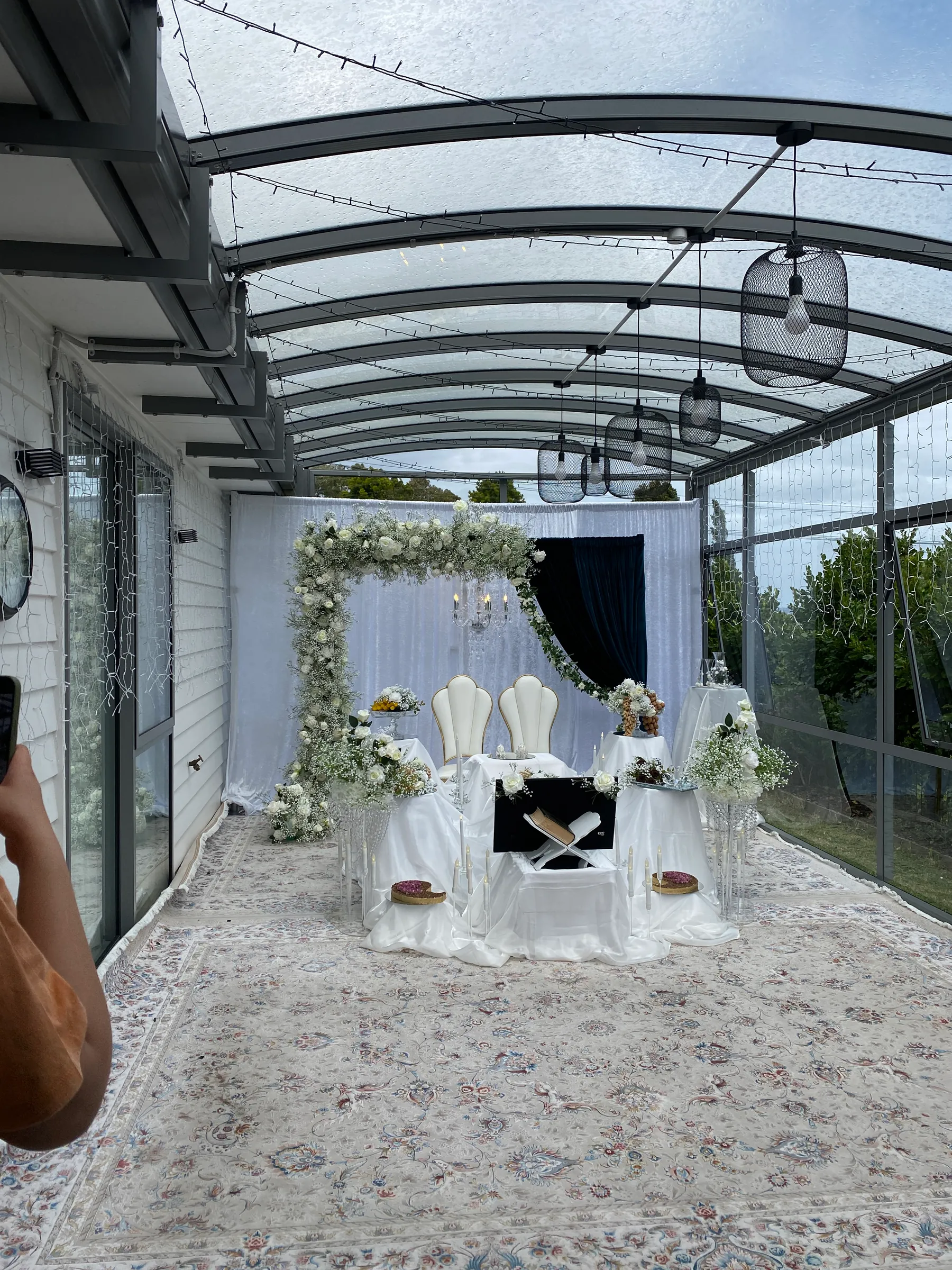 Cultural styling by Decoration X: Sofreh aghd on a glass-roofed patio: white satin spread, Quran stand and mirror, two shell chairs under a white floral arch, Persian rug and pendant lights.