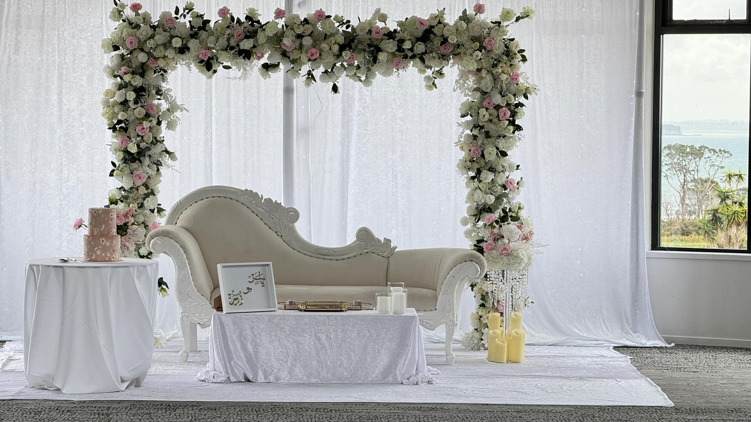 Stage styling by Decoration X: White carved chaise under a square arch of pink and ivory roses, a two-tier blush cake, a low table with a framed plaque and candles, sea view out the window.
