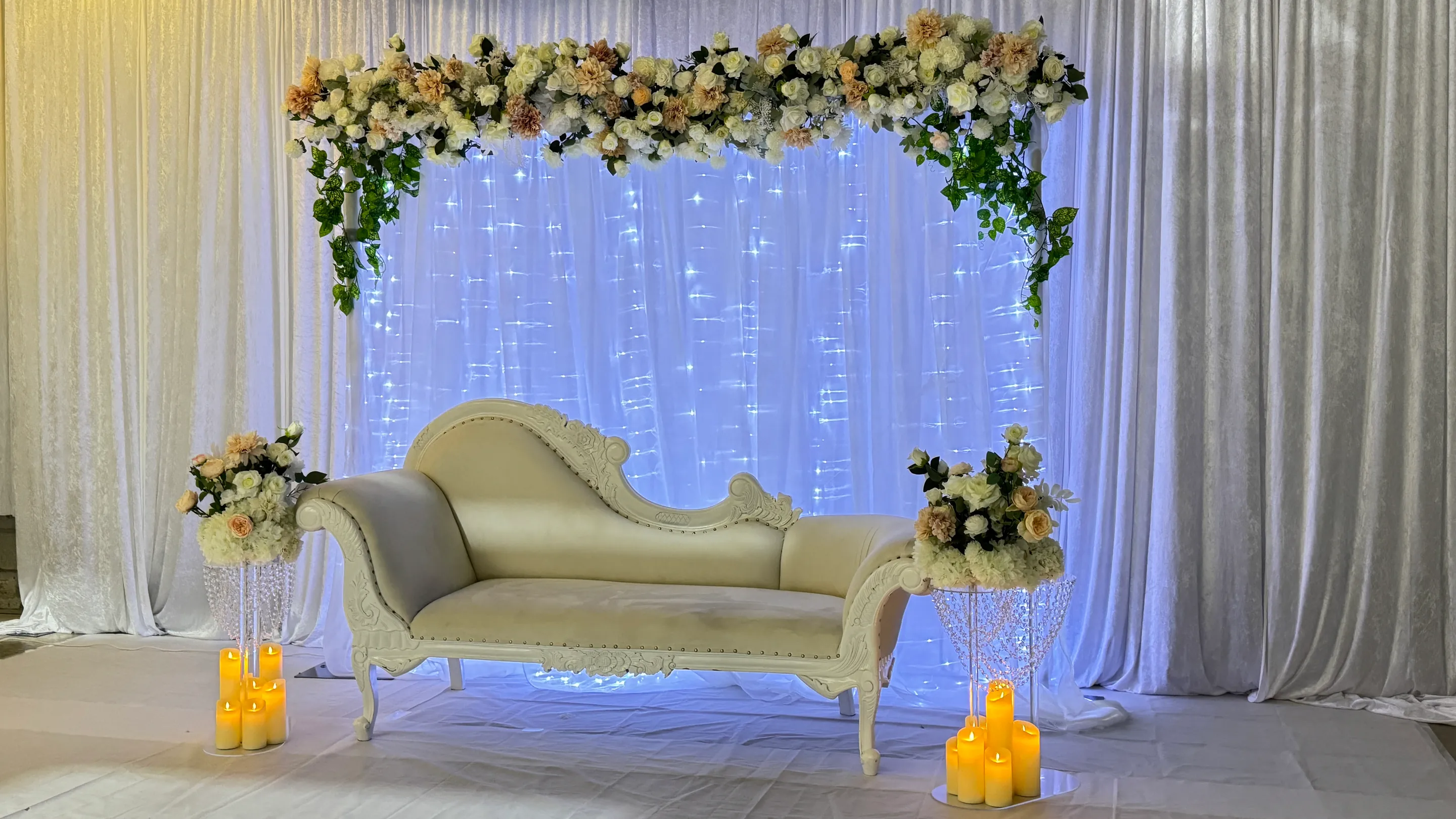 Stage styling by Decoration X: White carved chaise under a garland of peach and ivory roses with trailing ivy, a fairy-lit sheer panel behind, crystal posy stands and pillar candles.