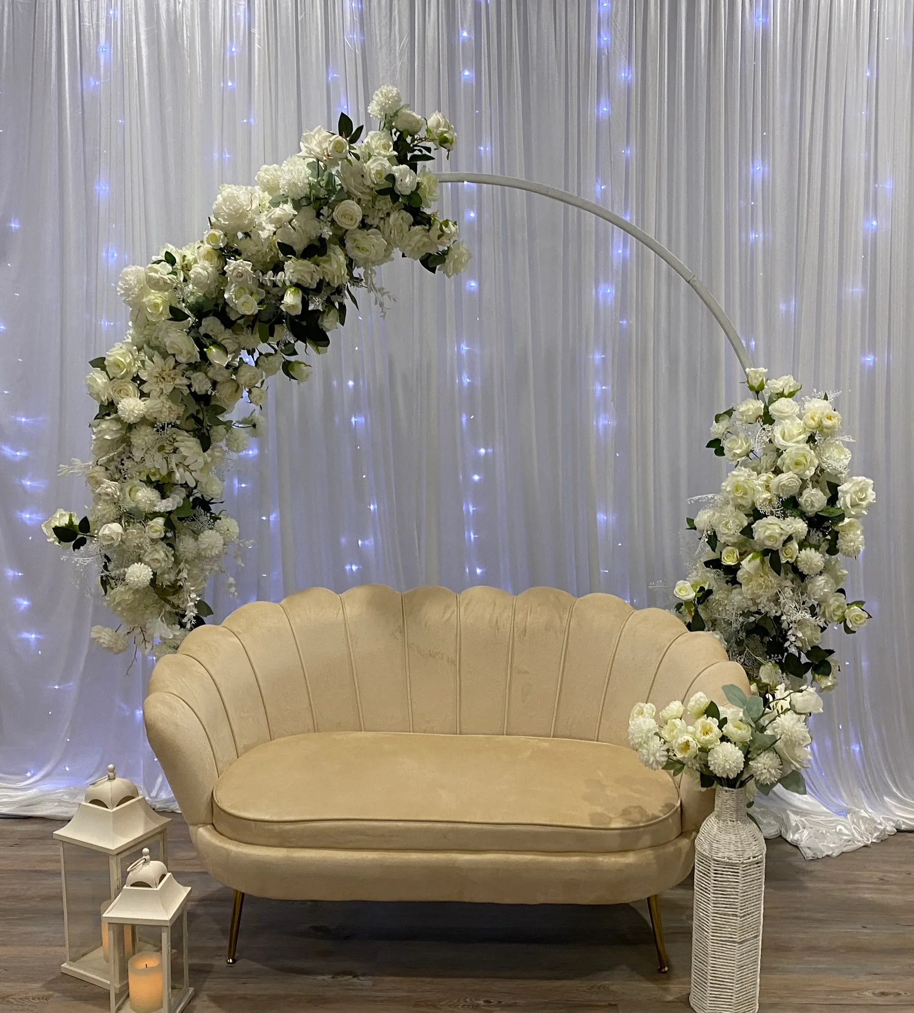 Stage styling by Decoration X: Cream scalloped velvet loveseat under a white hoop arch of ivory roses and gypsophila, a fairy-lit sheer drape behind, white lanterns and a wicker vase.