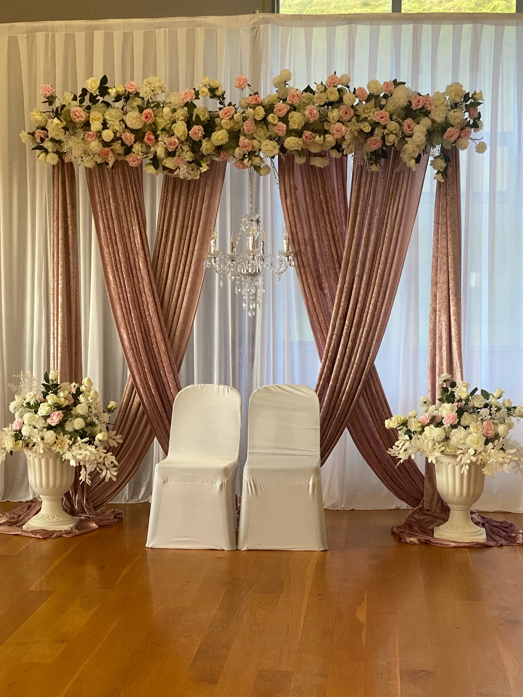 Stage styling by Decoration X: Two covered chairs before crossed dusty-pink velvet drapes and white sheers, a pink and cream rose garland above, a crystal chandelier and urns of flowers.