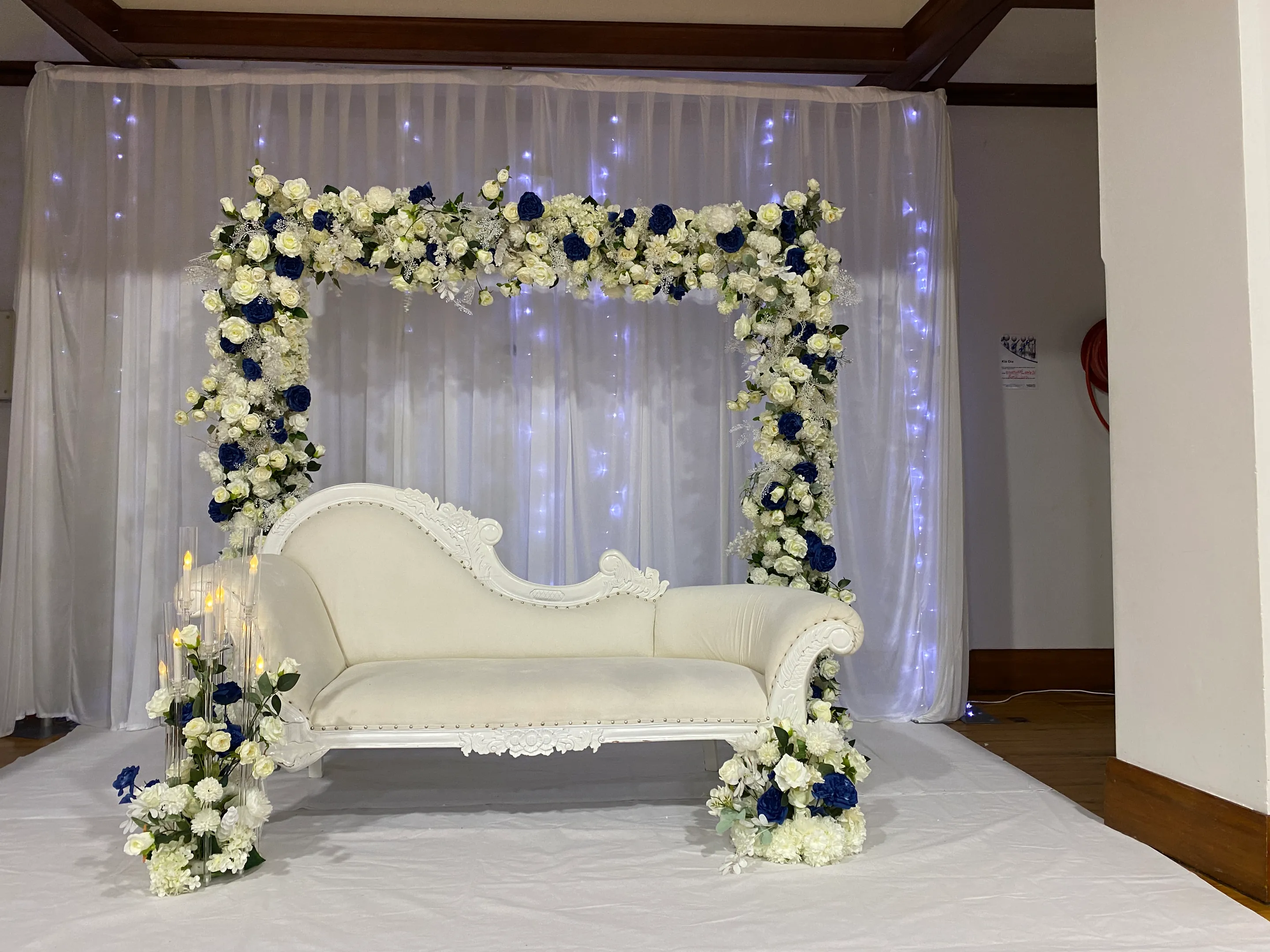 Stage styling by Decoration X: White carved chaise beneath a square arch of white and navy roses, a fairy-lit sheer drape behind and candle cylinders with florals at its foot.