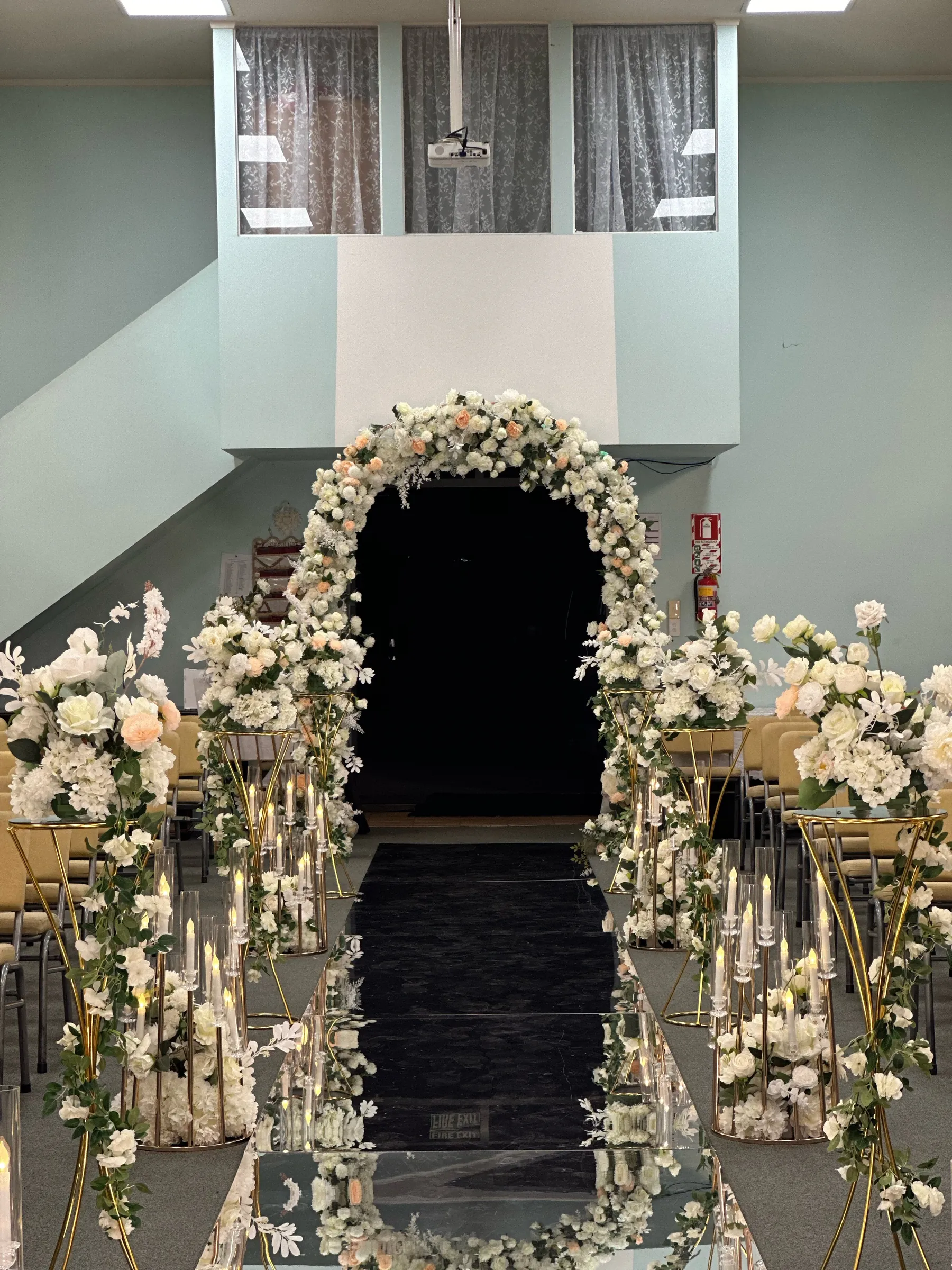 Ceremony styling by Decoration X: White and peach rose arch over a doorway, the black mirror aisle lined with gold stands of white roses and clear candle cylinders bedded in florals.