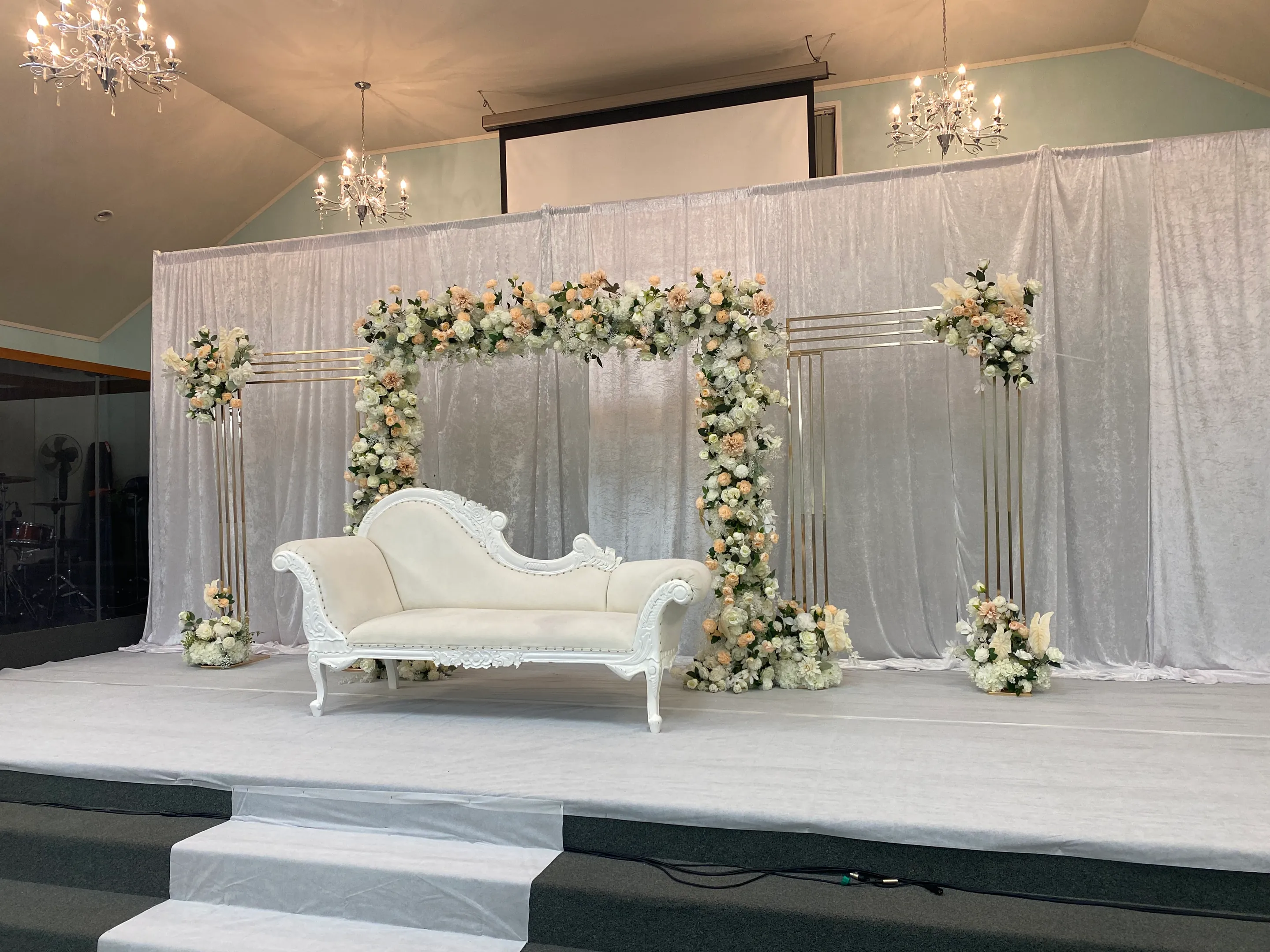 Stage styling by Decoration X: White carved chaise before a white velvet drape, framed by gold line panels and a square arch of peach and ivory roses, crystal chandeliers overhead.