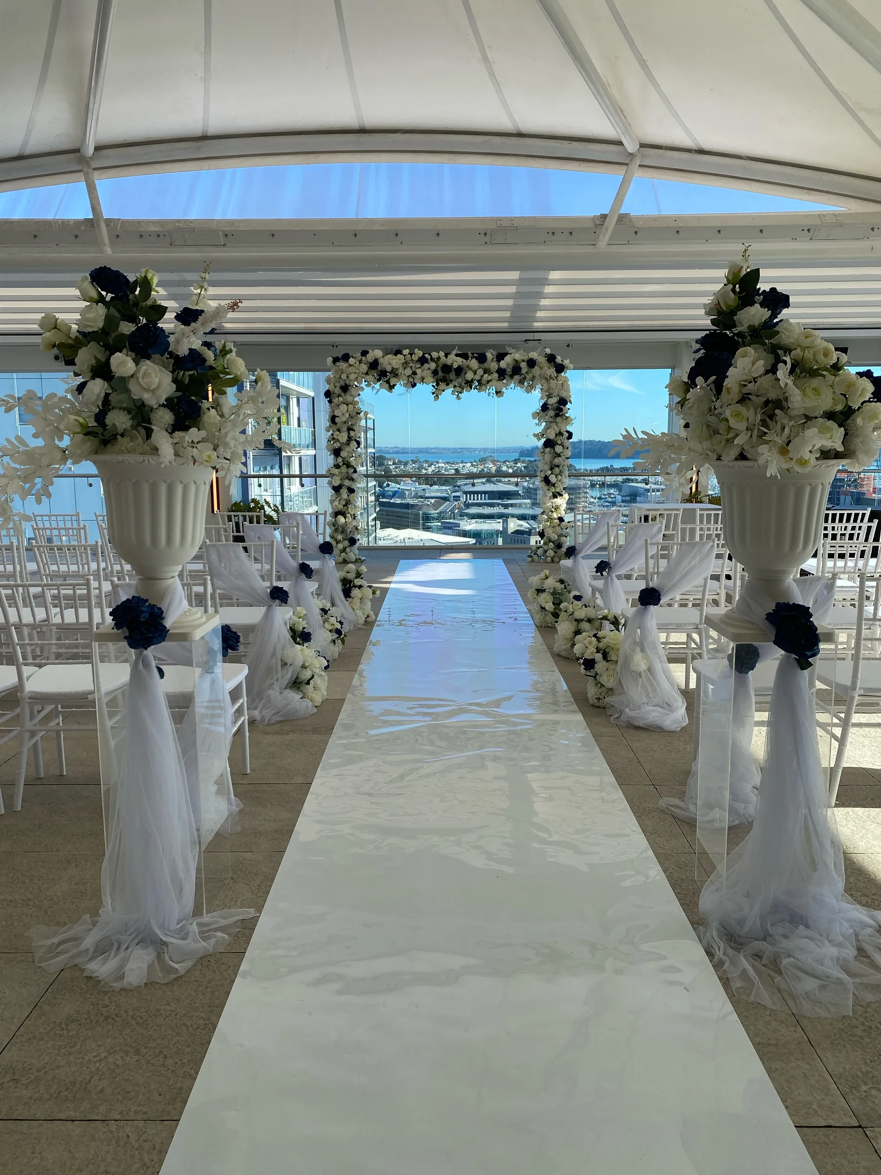 Ceremony styling by Decoration X: Rooftop ceremony aisle: white gloss runner between tulle-tied white chairs, urns of white and navy roses on acrylic plinths, a floral arch framing the harbour.
