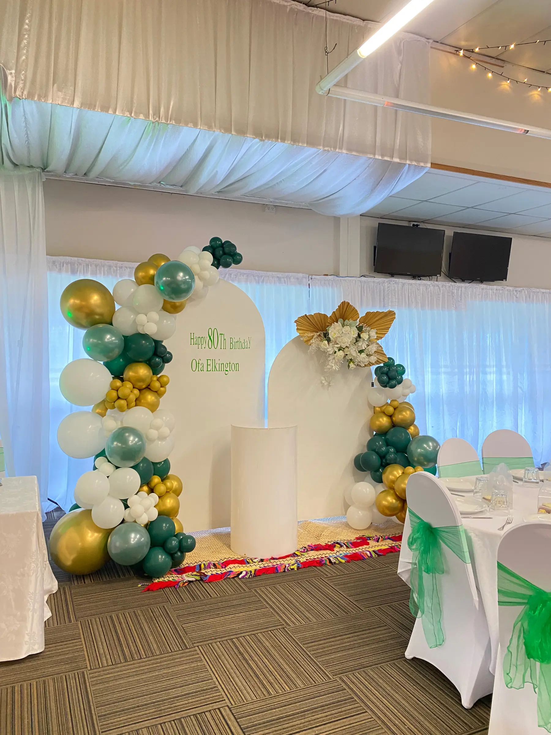 Birthday styling by Decoration X: 'Happy 80th Birthday Ofa Elkington' green/gold balloons, arch boards; TVs on wall.