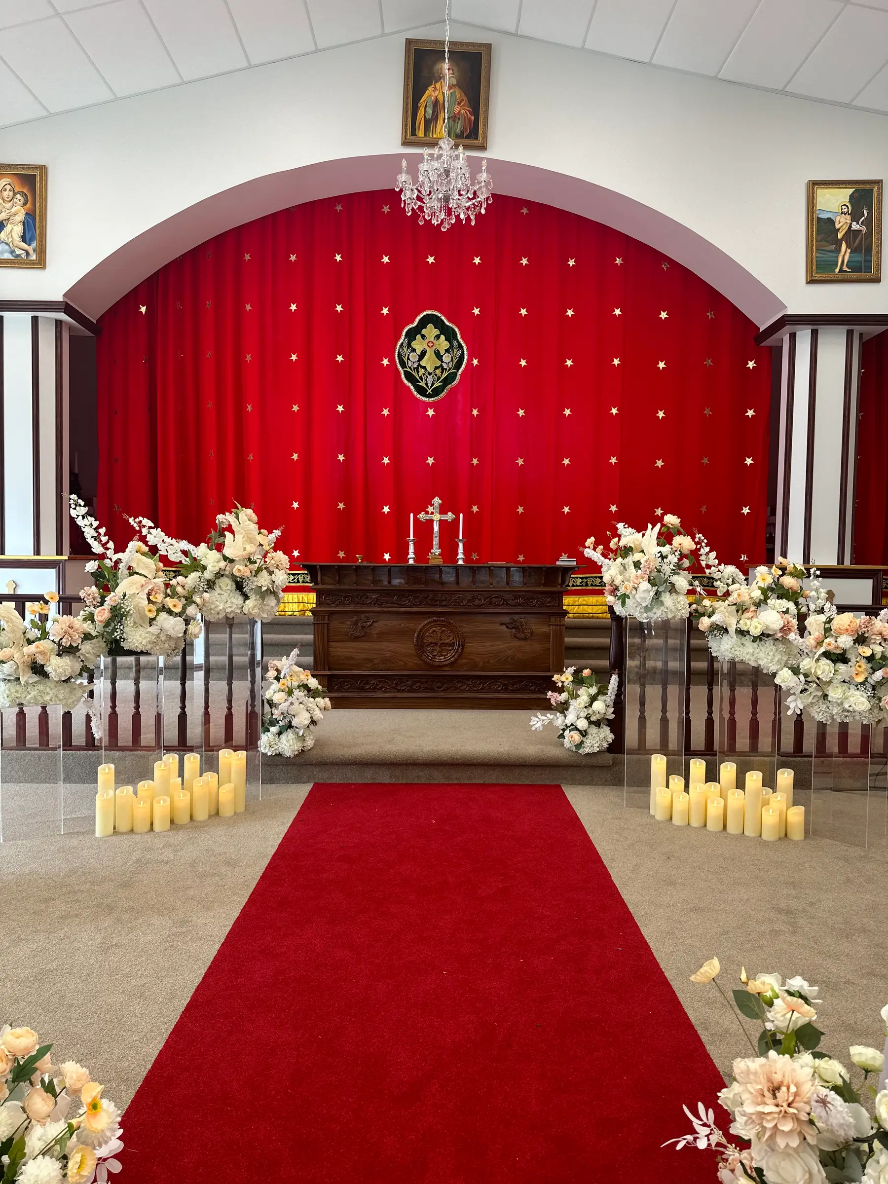 Cultural styling by Decoration X: Church aisle laid with red carpet between white and peach florals on acrylic plinths, pillar candles, a gold-starred red drape behind the altar.