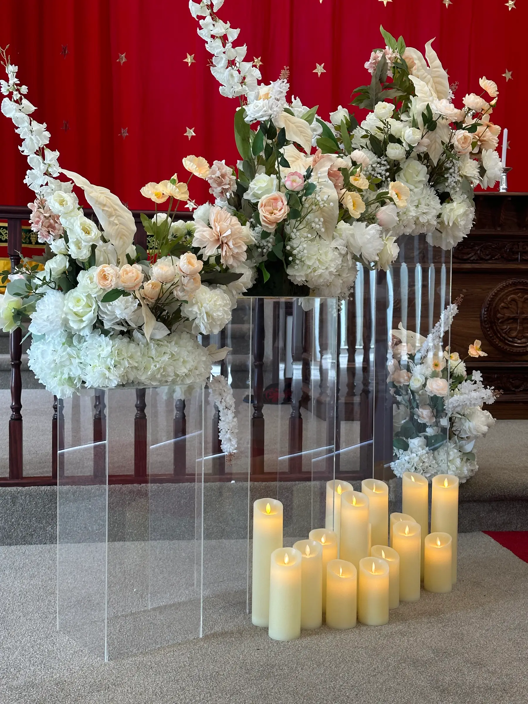 Cultural styling by Decoration X: Tall acrylic plinths carrying white hydrangea and peach roses beside a bank of pillar candles, gold-starred red drape behind.