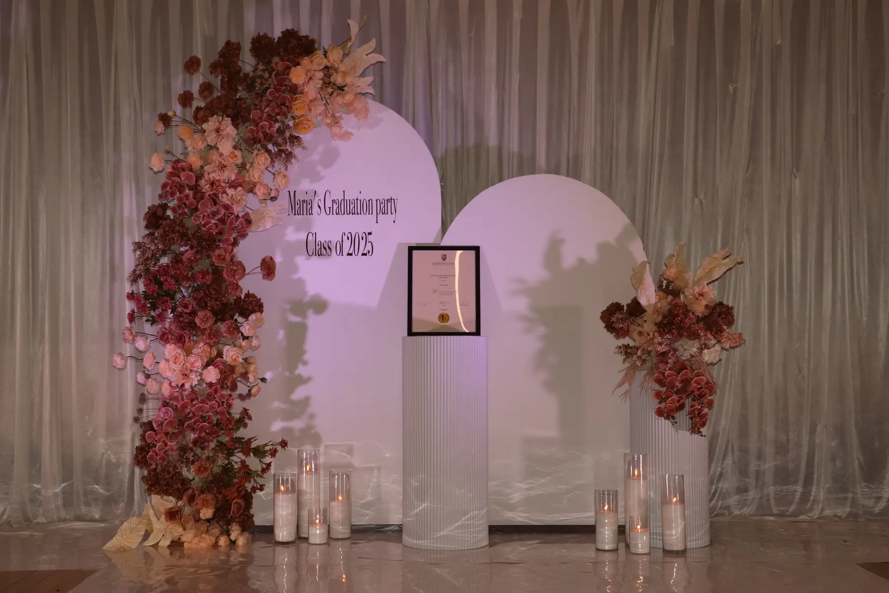 Graduation styling by Decoration X: Burgundy and blush floral arch over a white arched backdrop, fluted plinths and pillar candles on a mirrored floor.