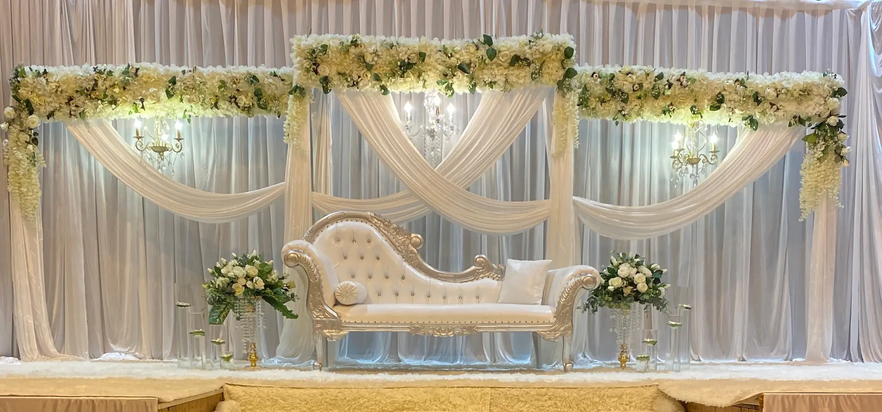 Stage styling by Decoration X: Silver chaise on stage, triple drape swags, chandeliers, floral garlands.