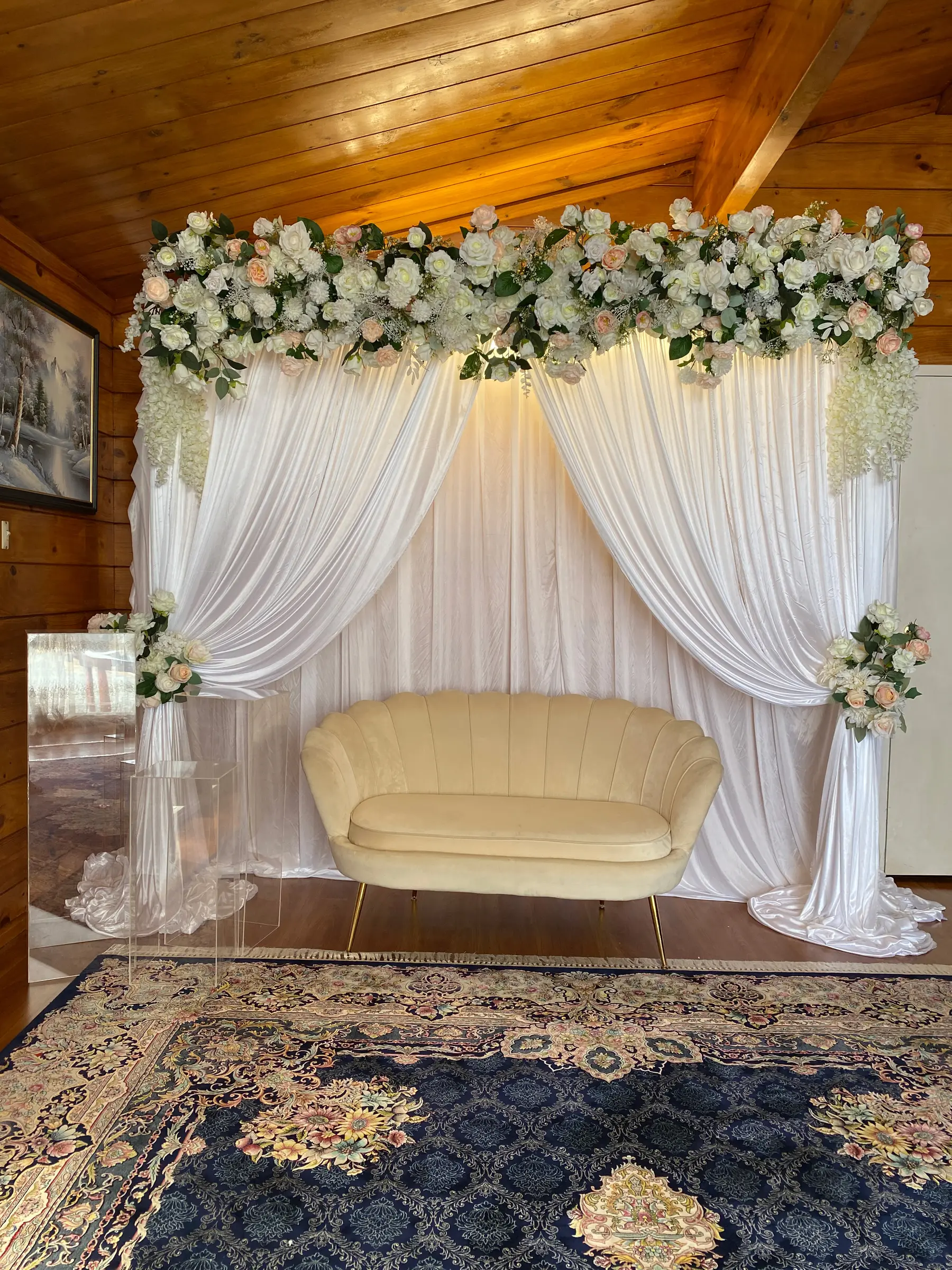 Stage styling by Decoration X: Cream scallop loveseat under drapes and garland, timber ceiling, Persian rug.