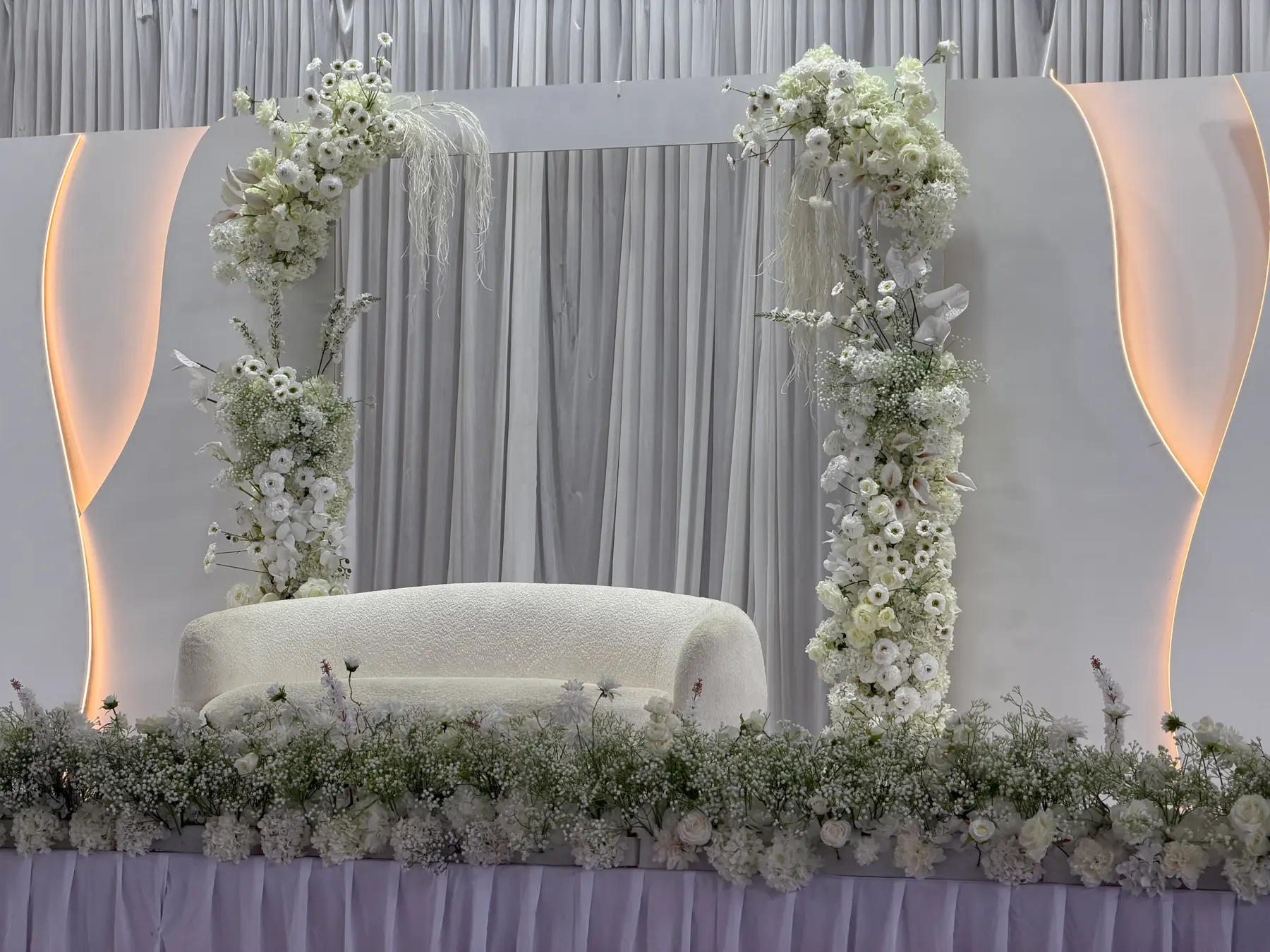 Stage styling by Decoration X: White boucle loveseat on a stage between two white floral columns, gypsophila runner along the front, warm cove lighting behind.