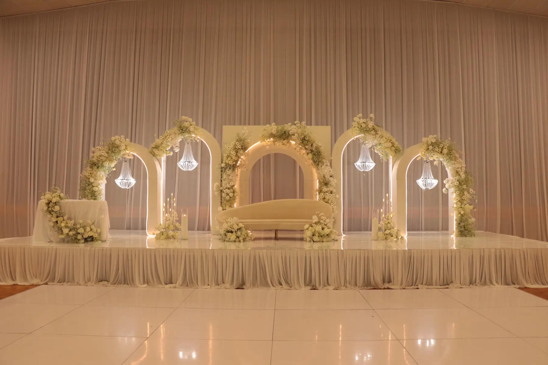 Stage styling by Decoration X: Five illuminated arches with crystal chandeliers and white florals above a cream sofa, draped stage skirt and a gloss dance floor.