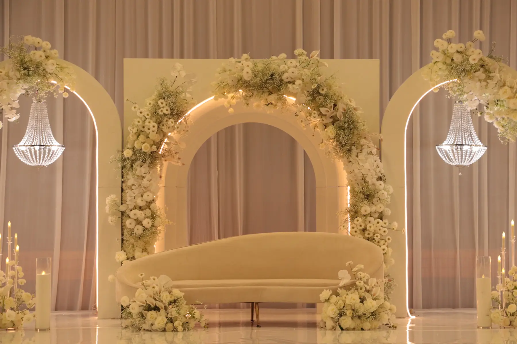 Stage styling by Decoration X: Cream sofa beneath a lit arch heavy with white ranunculus and gypsophila, a crystal chandelier either side.