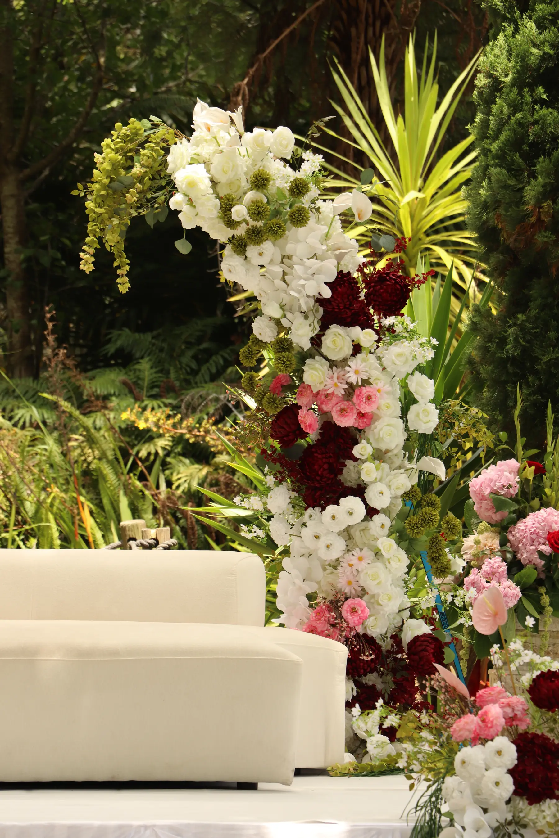 Stage styling by Decoration X: Cream sofas on an outdoor stage beside a floral column of burgundy dahlias, pink roses and white orchids, against native bush.