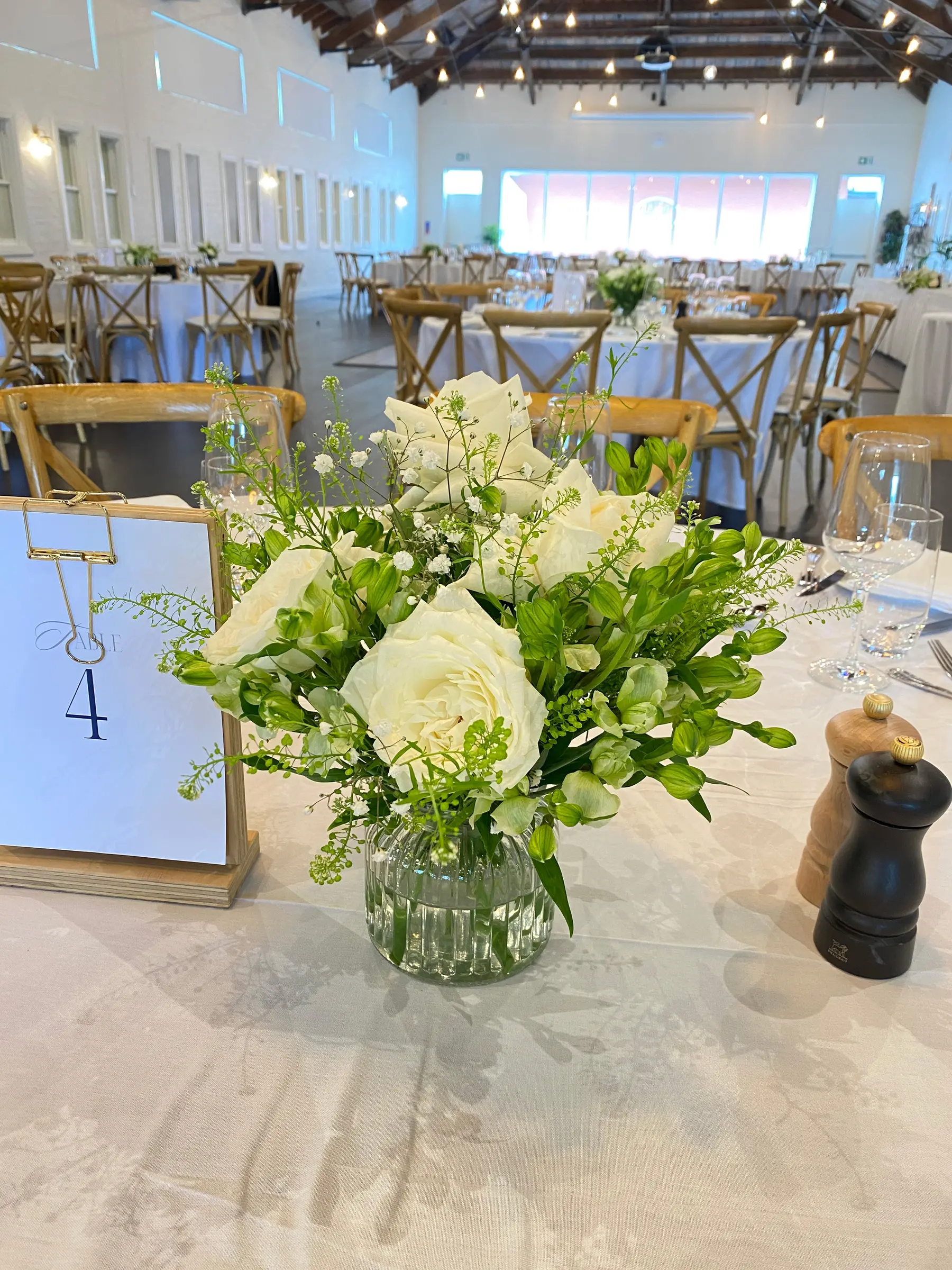 Tables styling by Decoration X: White rose centrepiece, 'Table 4' card, cross-back chairs, festoon-lit hall.
