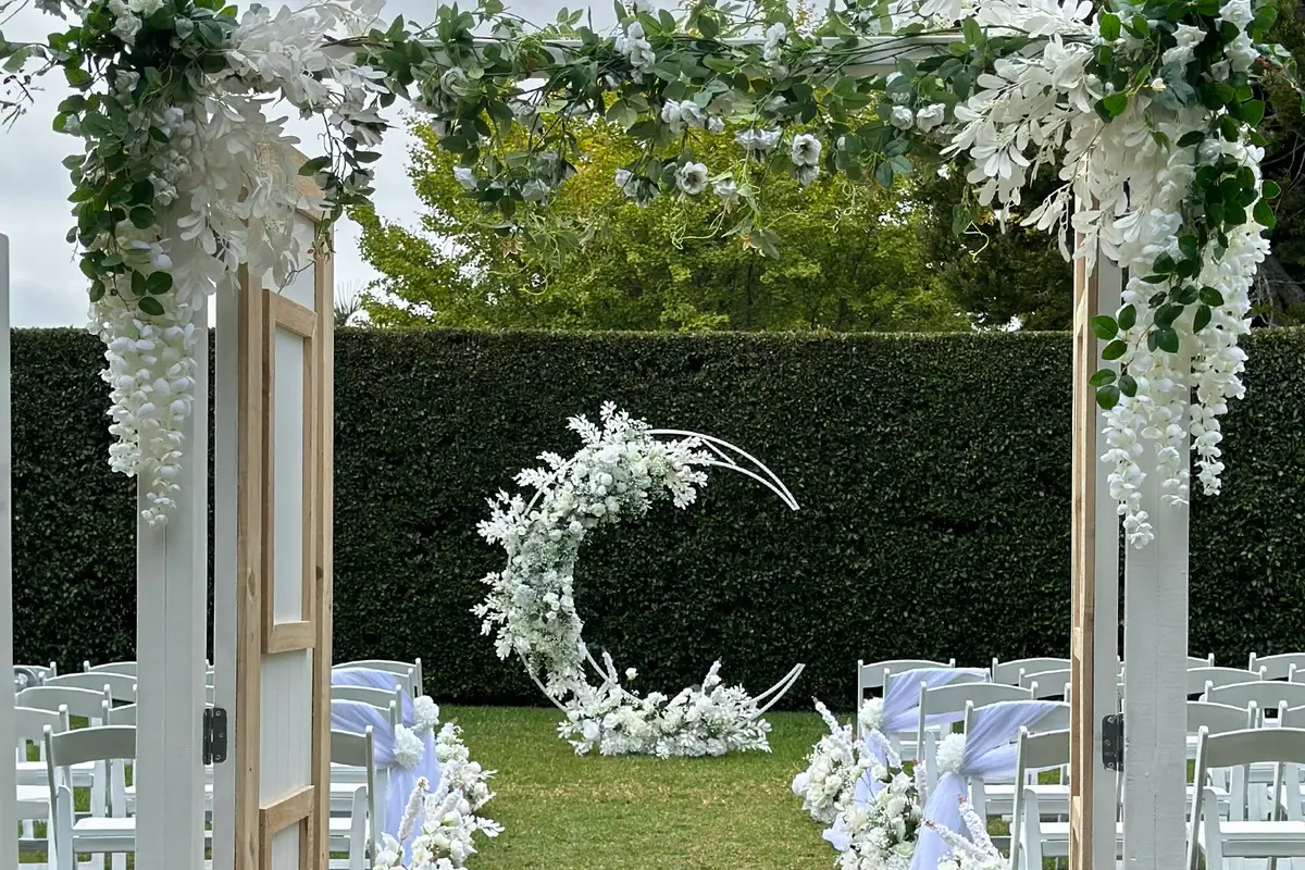 Ceremony styling by Decoration X: Moon arch on lawn framed through rustic doors; white chairs, sashes.