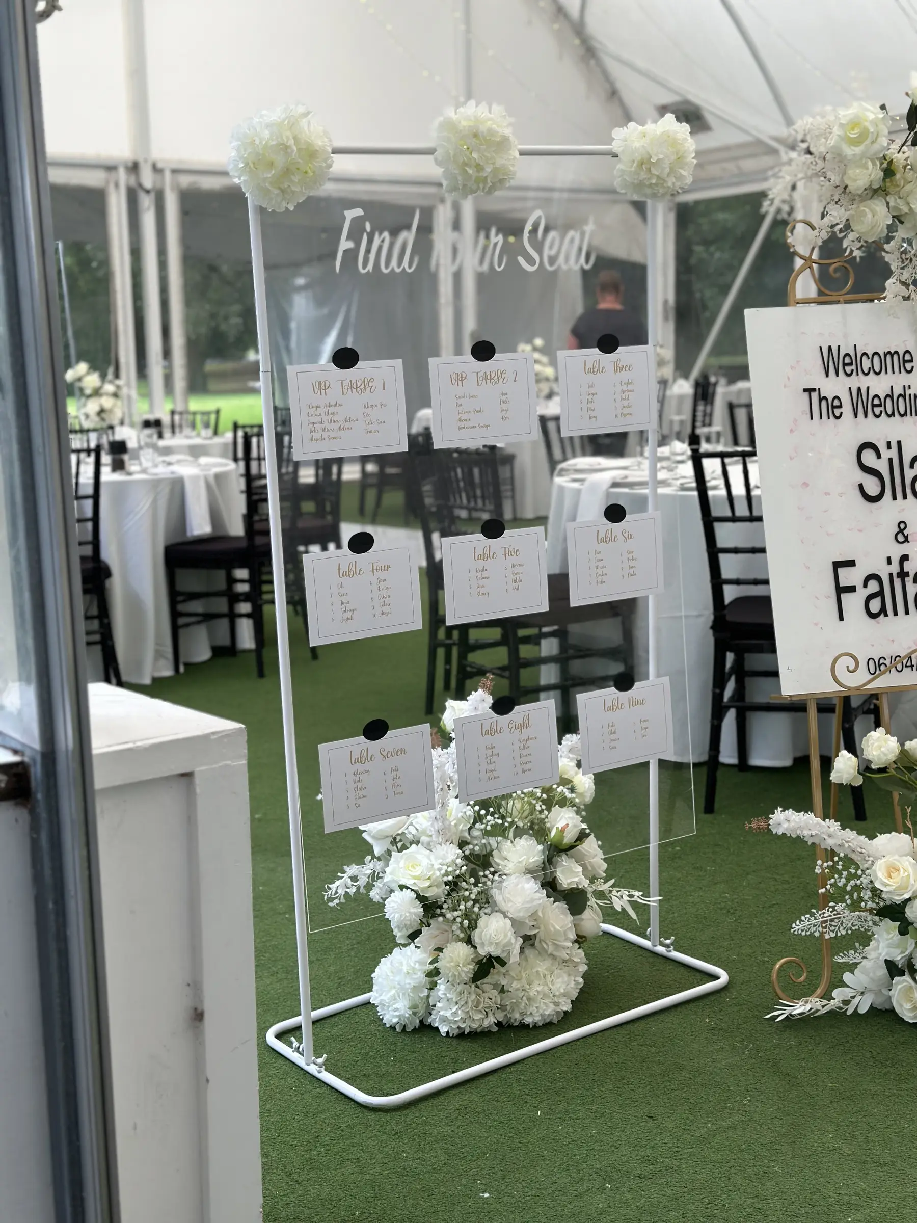 Ceremony styling by Decoration X: Welcome-sign easel with white florals inside marquee; tables behind.