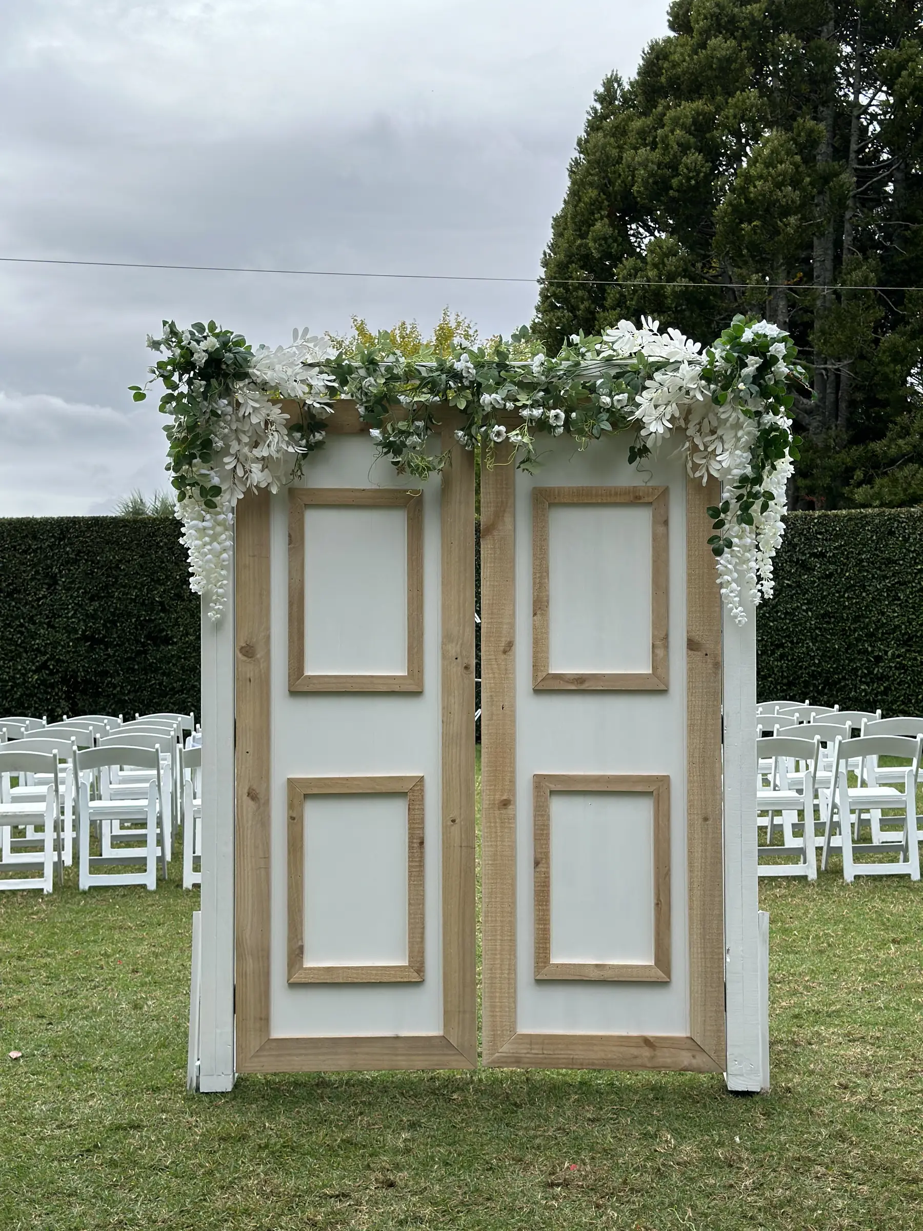 Ceremony styling by Decoration X: Rustic white doors arch with wisteria florals on lawn, white chairs.