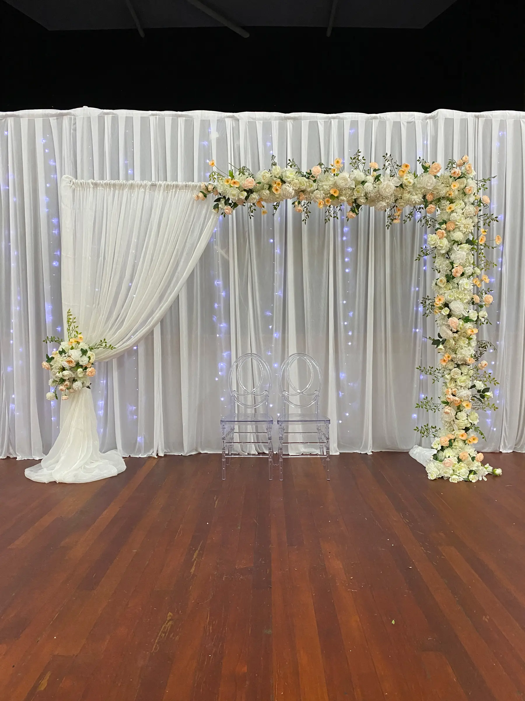 Ceremony styling by Decoration X: Peach floral L-garland on fairy-light drape, two ghost chairs; black ceiling.