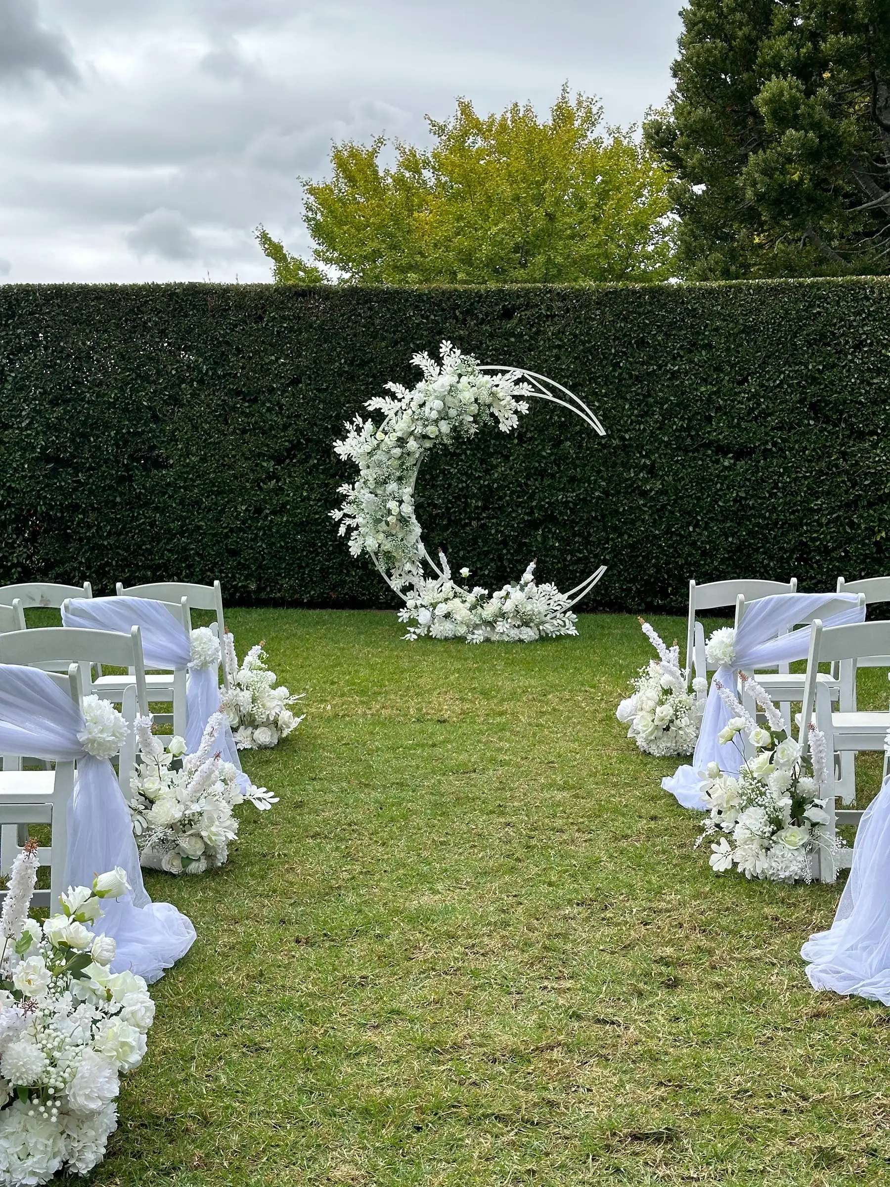 Ceremony styling by Decoration X: White floral moon arch on lawn, hedge, white chairs with sashes.