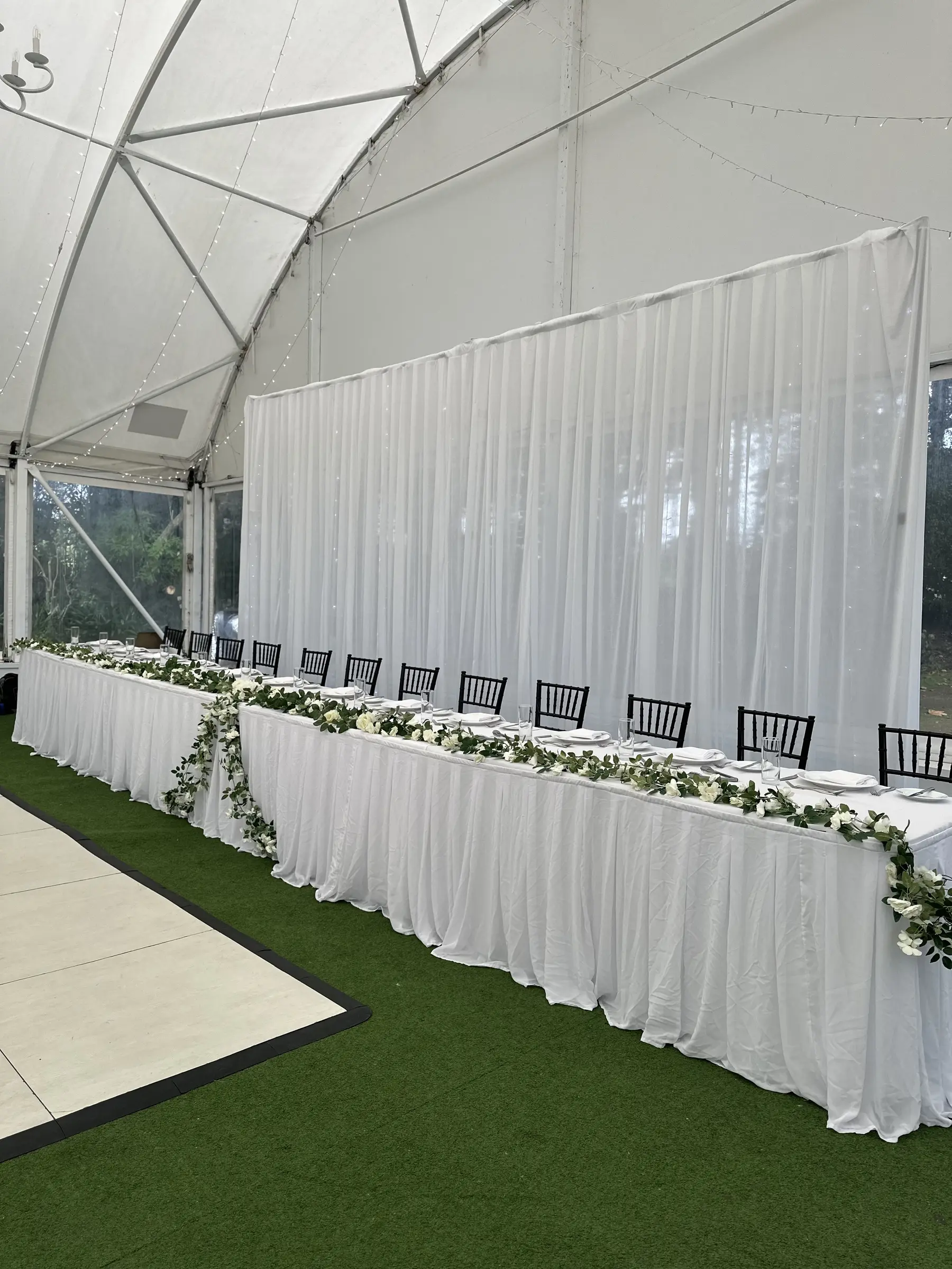 Reception styling by Decoration X: Marquee bridal table, white drape wall, greenery runner, black chiavari.