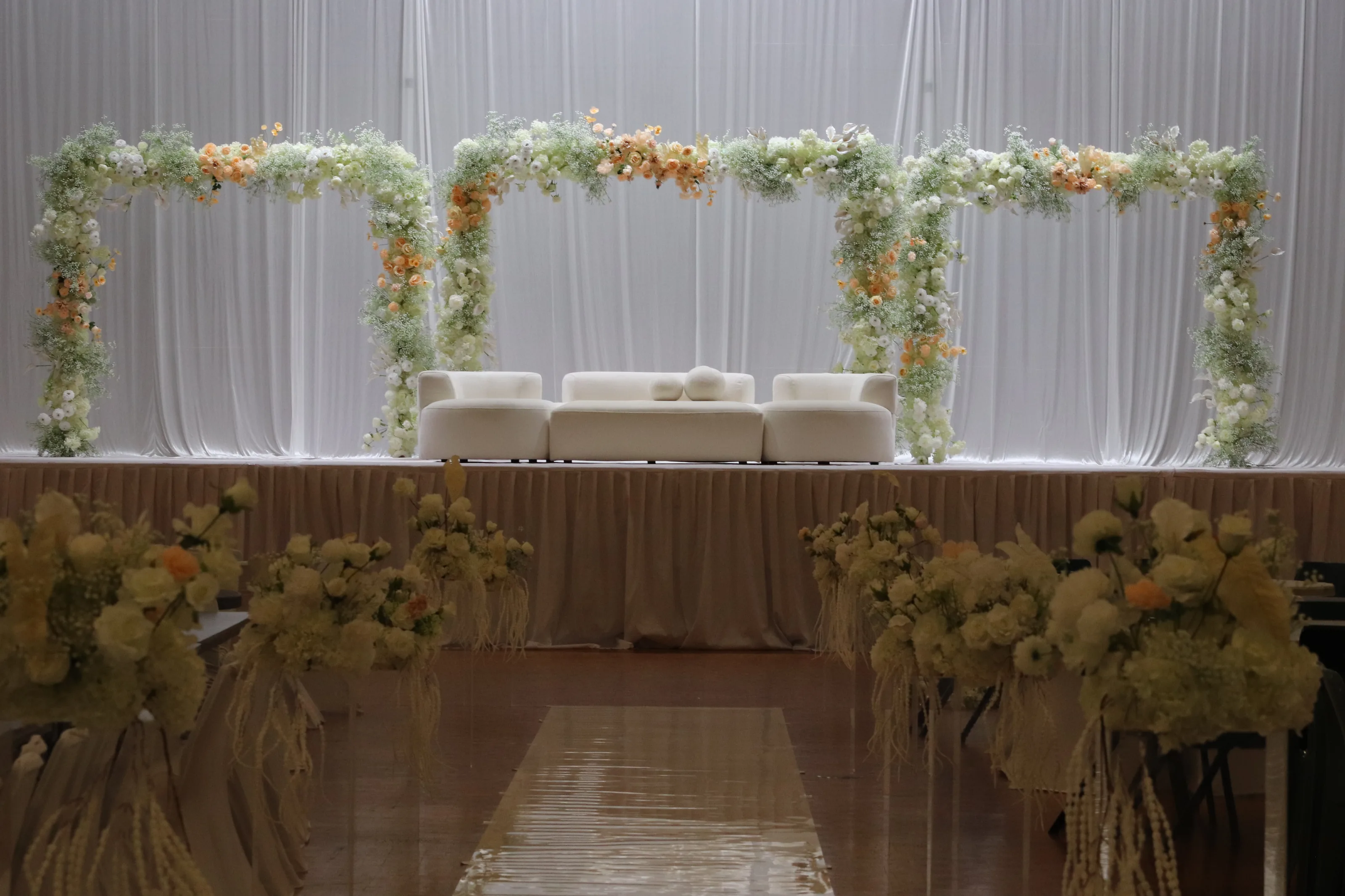 Reception styling by Decoration X: Three floral arches spanning a draped stage above white modular sofas; aisle lined with cream and peach florals over a mirrored runner.